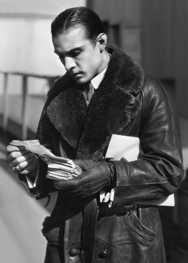 Rudolph Valentino in a fur-collared leather coat, reading a stack of letters, in a black-and-white portrait from the 1920s.