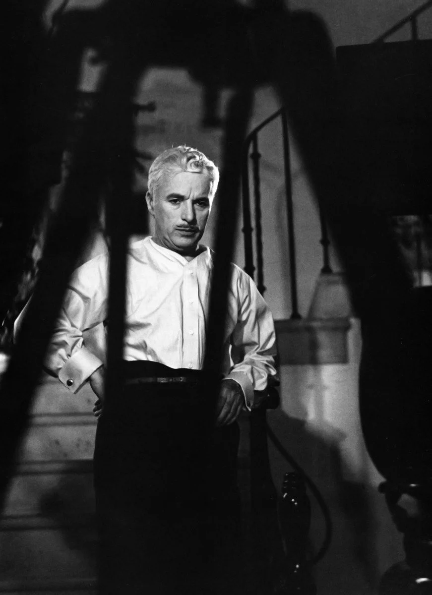 Charlie Chaplin, an older man with white hair and a thin mustache, stands on a staircase on the set of Monsieur Verdoux (1947), in a white shirt and dark trousers, one hand on his hip, seen through the bars of a banister.