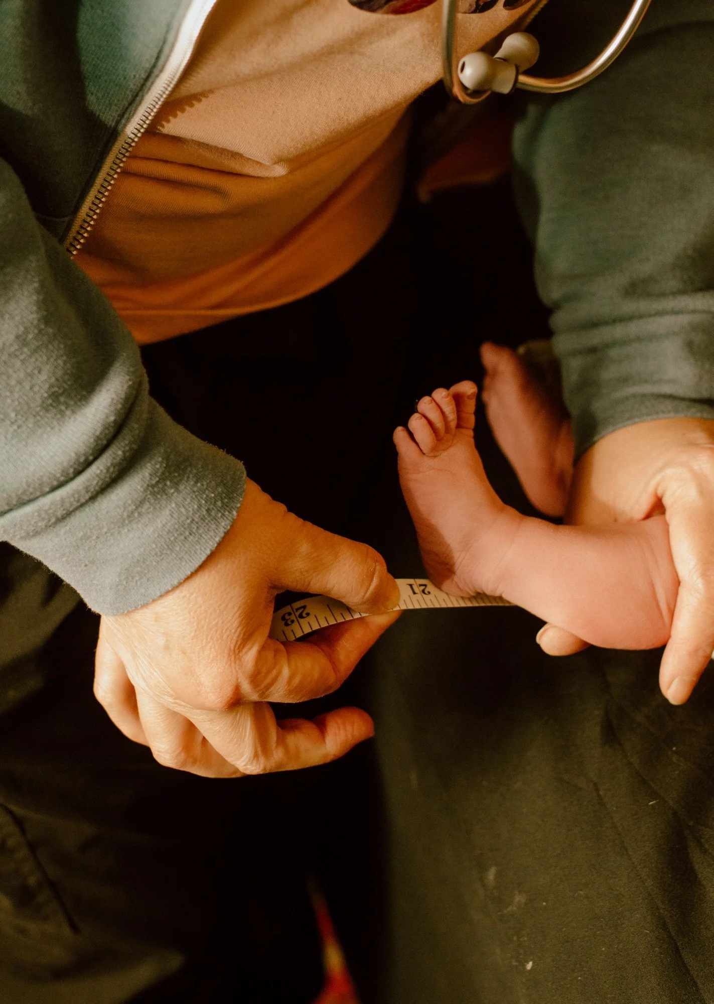 A close-up photo of an adult holding a measuring tape around a baby's tiny foot, with the adult wearing a gray sweatshirt and the baby having a small hand and foot with light skin. The background shows dark clothing and part of a wooden floor.