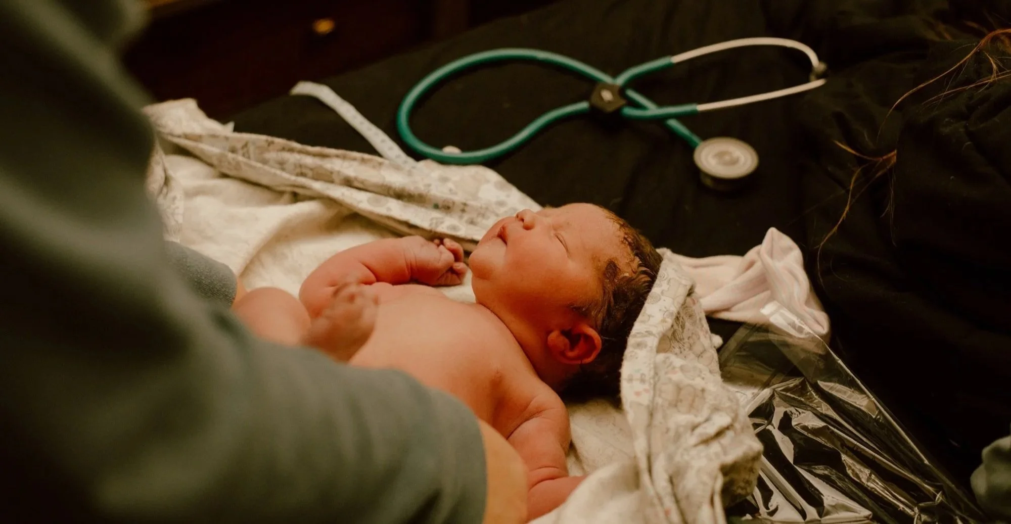 Newborn baby lying on a bed with a blanket, with a stethoscope placed on a black surface nearby, in a homebirth or birthing center setting.