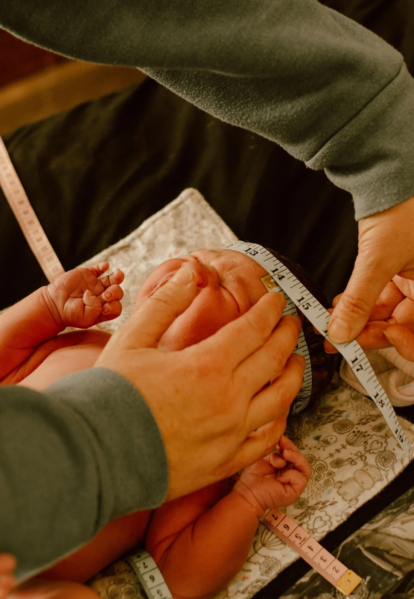 Close-up of a newborn baby being measured with a measuring tape by adults, with the baby's head and tiny hand visible. The scene is set on a patterned cloth.