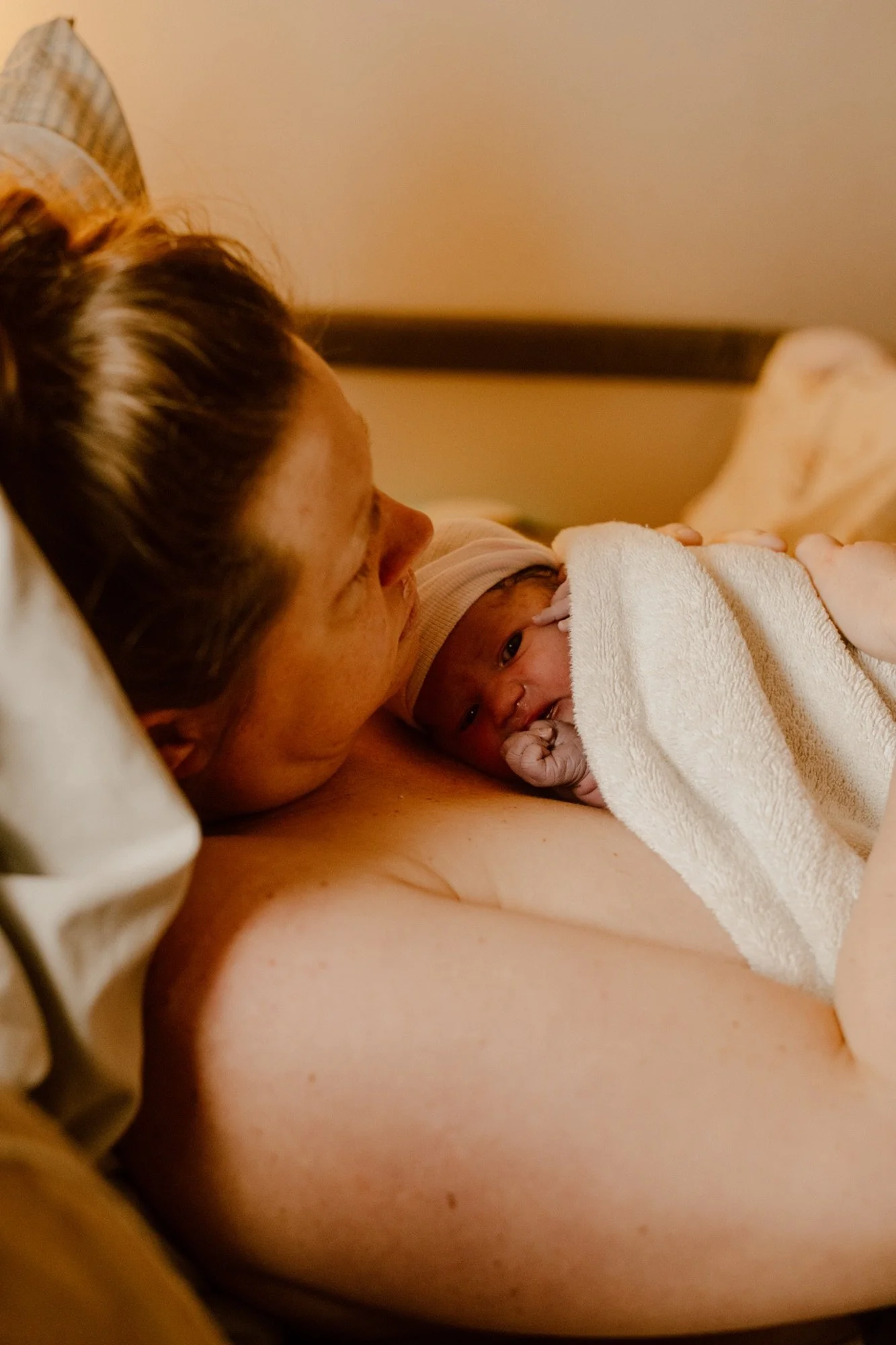 A person lying in bed holding a newborn baby wrapped in a white towel or blanket, with the person resting their head on a pillow and looking at the baby. Comfortable birth center and homebirth care.