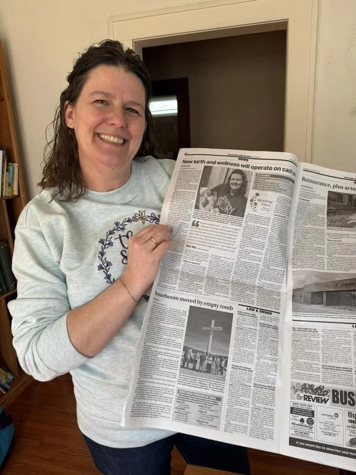 A woman with shoulder-length dark hair smiling and holding a newspaper in an indoor setting.