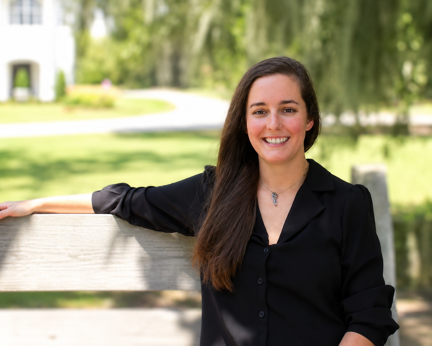 A woman with long brown hair, wearing a black blouse and a silver necklace, leaning on a wooden bench outdoors with a blurred background of trees and grass, smiling at the camera.