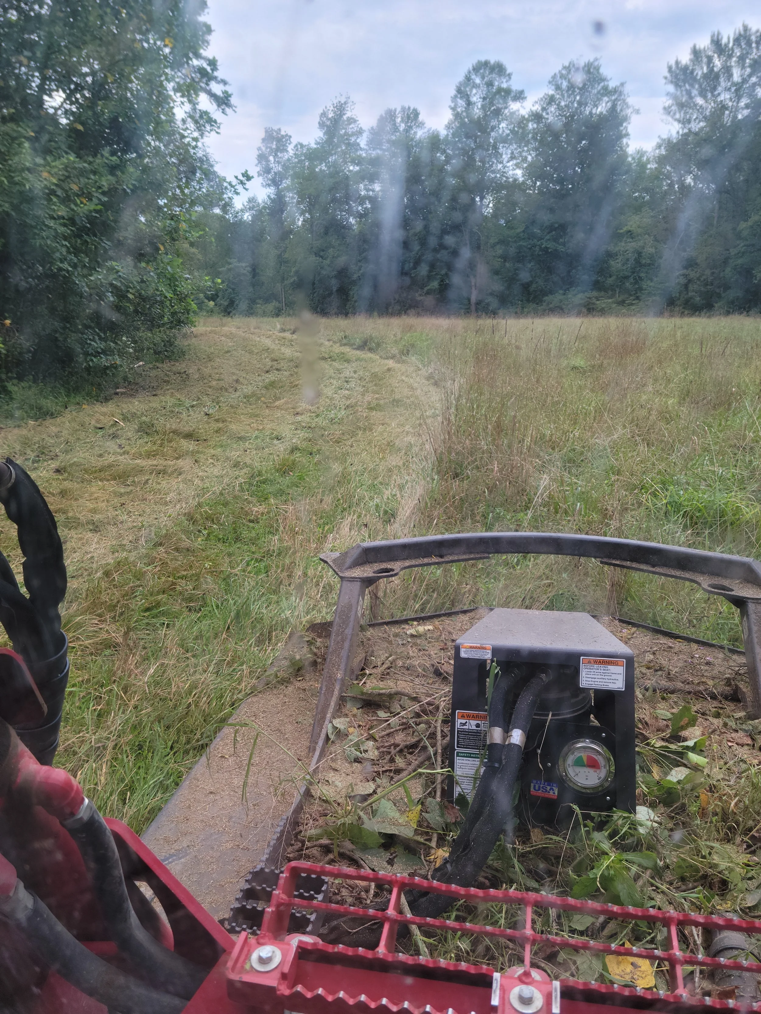 View from the front of a Skid Steer, Brush hogging a Grass pathway in a grassy field with trees in the background, on a cloudy day.   Delaware Grading Services