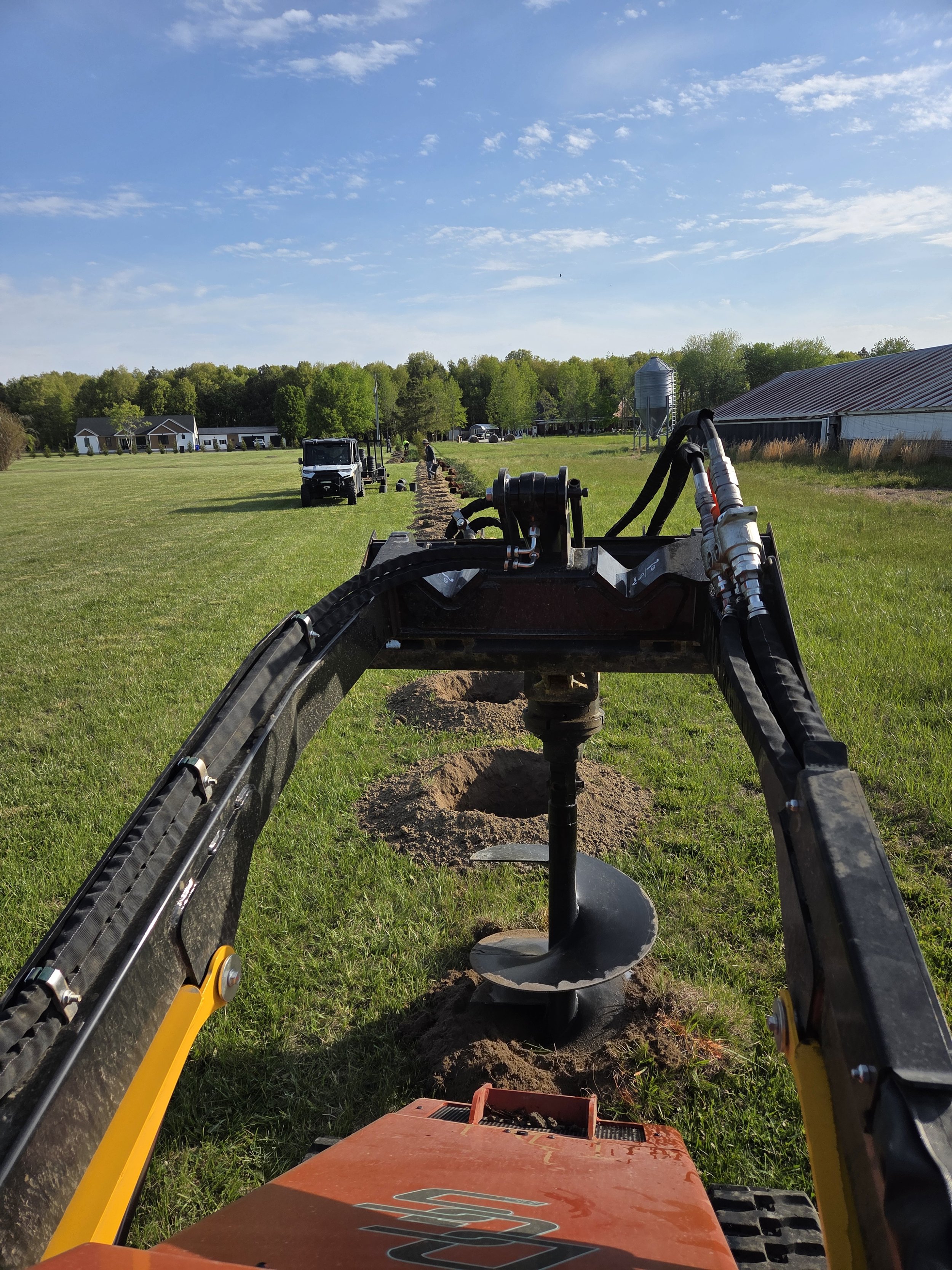 A view from a construction or excavation vehicle showing a row of holes being drilled into the ground in a grassy field. Several workers from  Delaware Grading Services are visible in the background, planting small trees into the holes