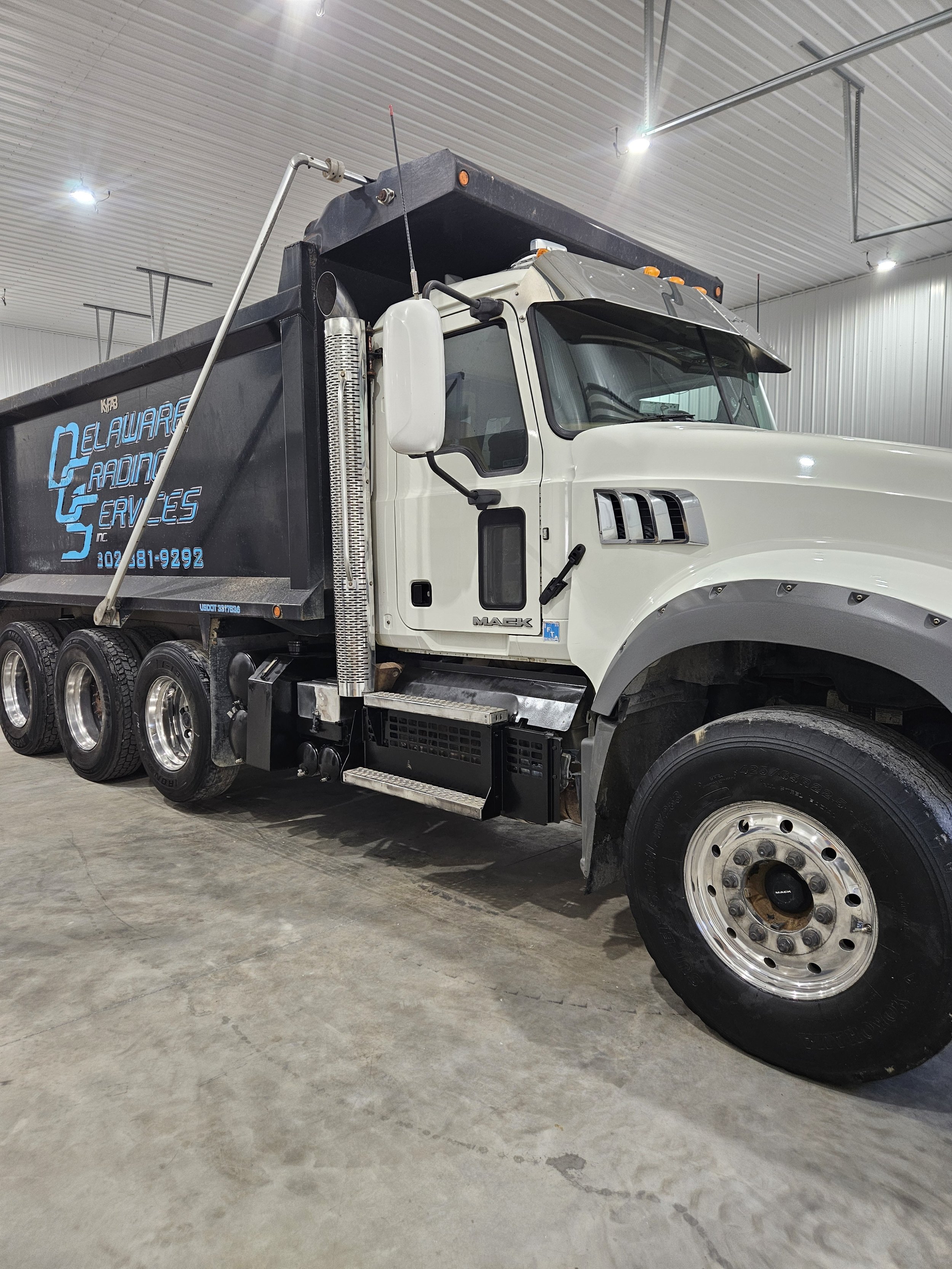 A white Mack truck with a black cargo bed, parked inside a warehouse with metal walls and ceiling. The truck has multiple wheels and a large fuel tank visible on the side. Showcasing Delaware Grading services ability to haul material