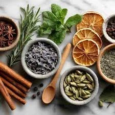 A variety of spices and herbs on a light surface, including cinnamon sticks, star anise, dried citrus slices, black and green cardamom, and dried lavender, with some in small bowls and others scattered.