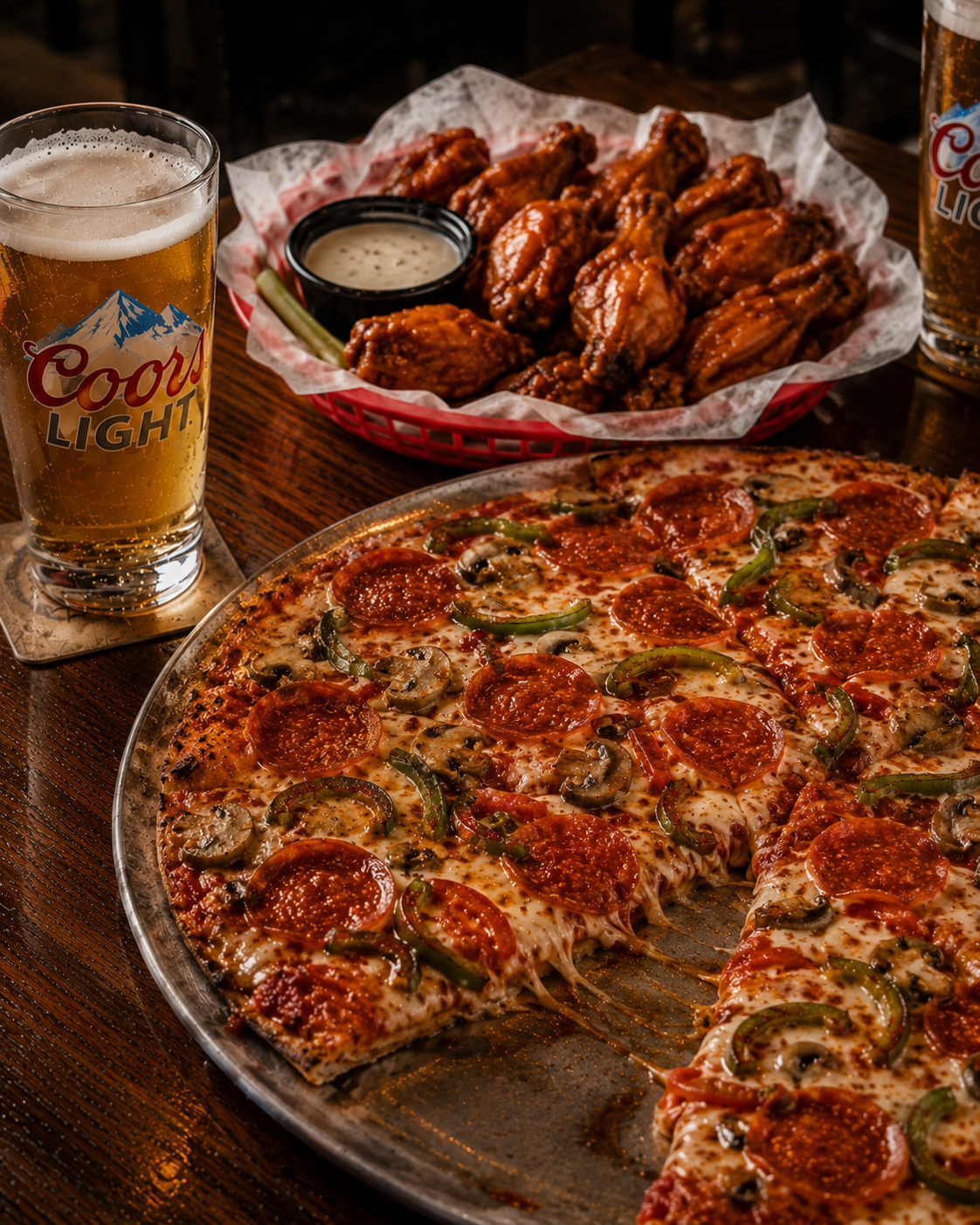 A pizza with pepperoni, green peppers, and mushrooms on a metal tray, a basket of chicken wings with ranch dipping sauce, and two glasses of Coors Light beer on a wooden table.