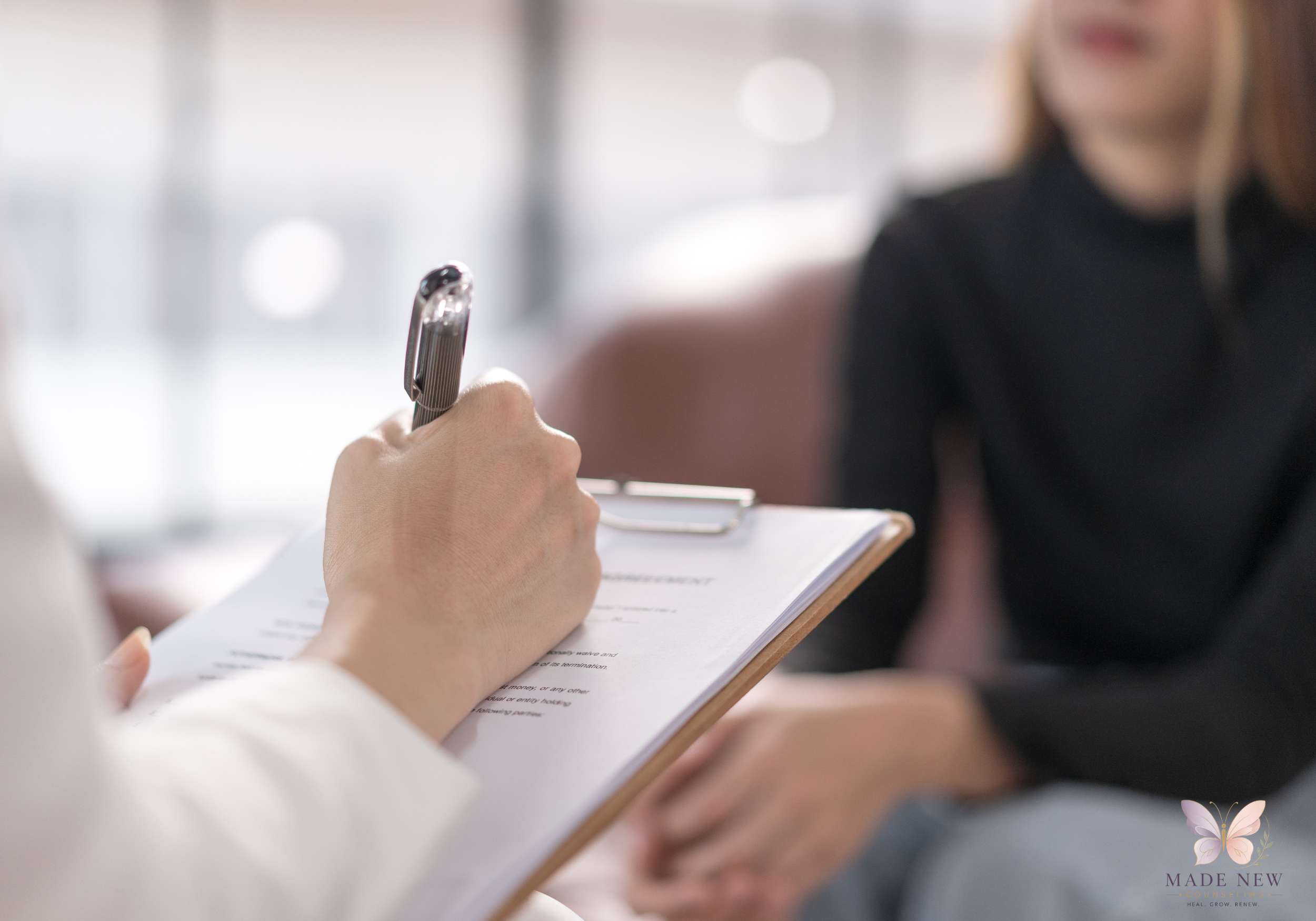 Close-up of a therapist’s hand writing on a clipboard labeled “Assessment” while a client sits blurred in the background.