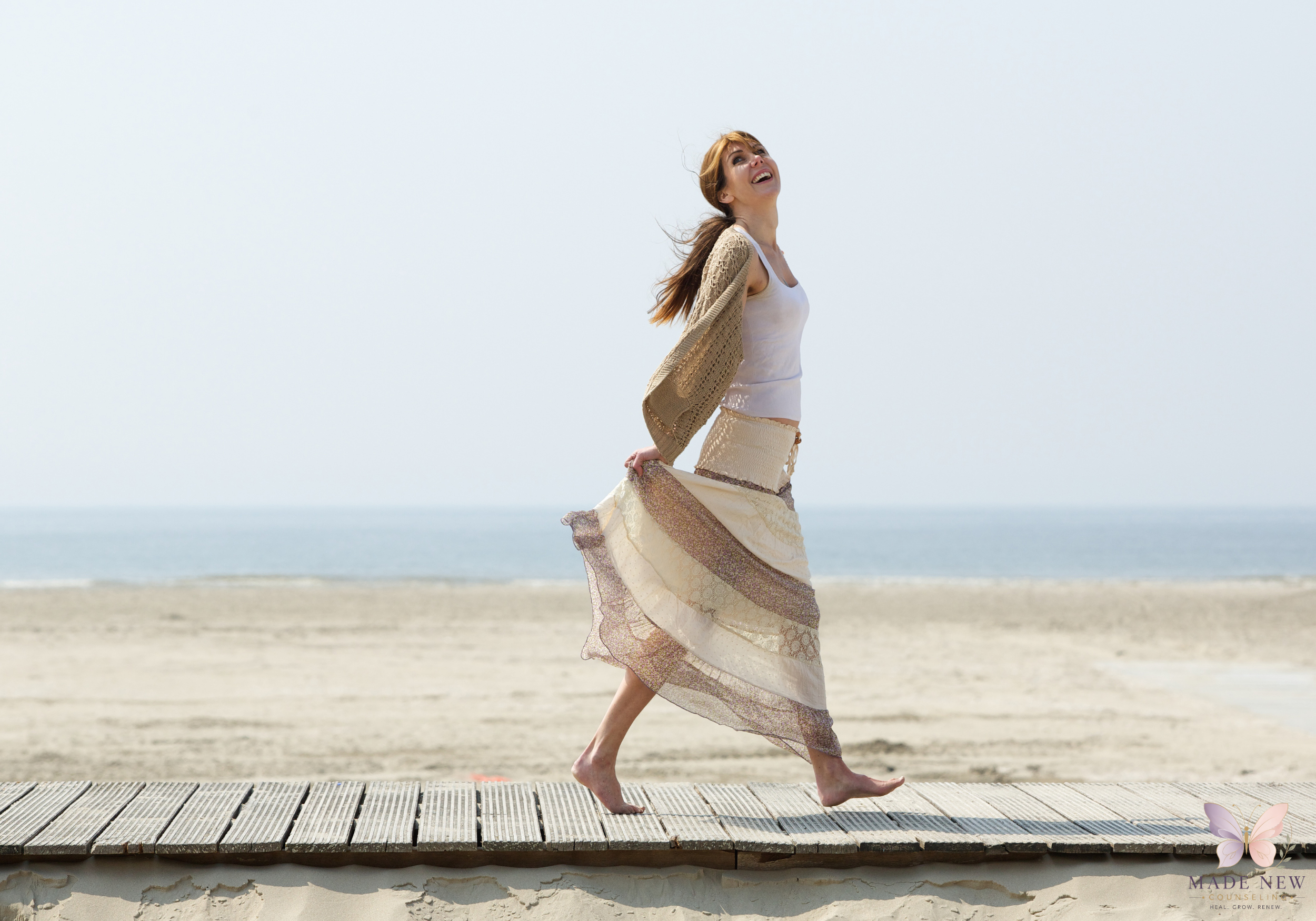 Joyful woman runs barefoot along a wooden beach boardwalk, arms slightly out, smiling up at the sky.