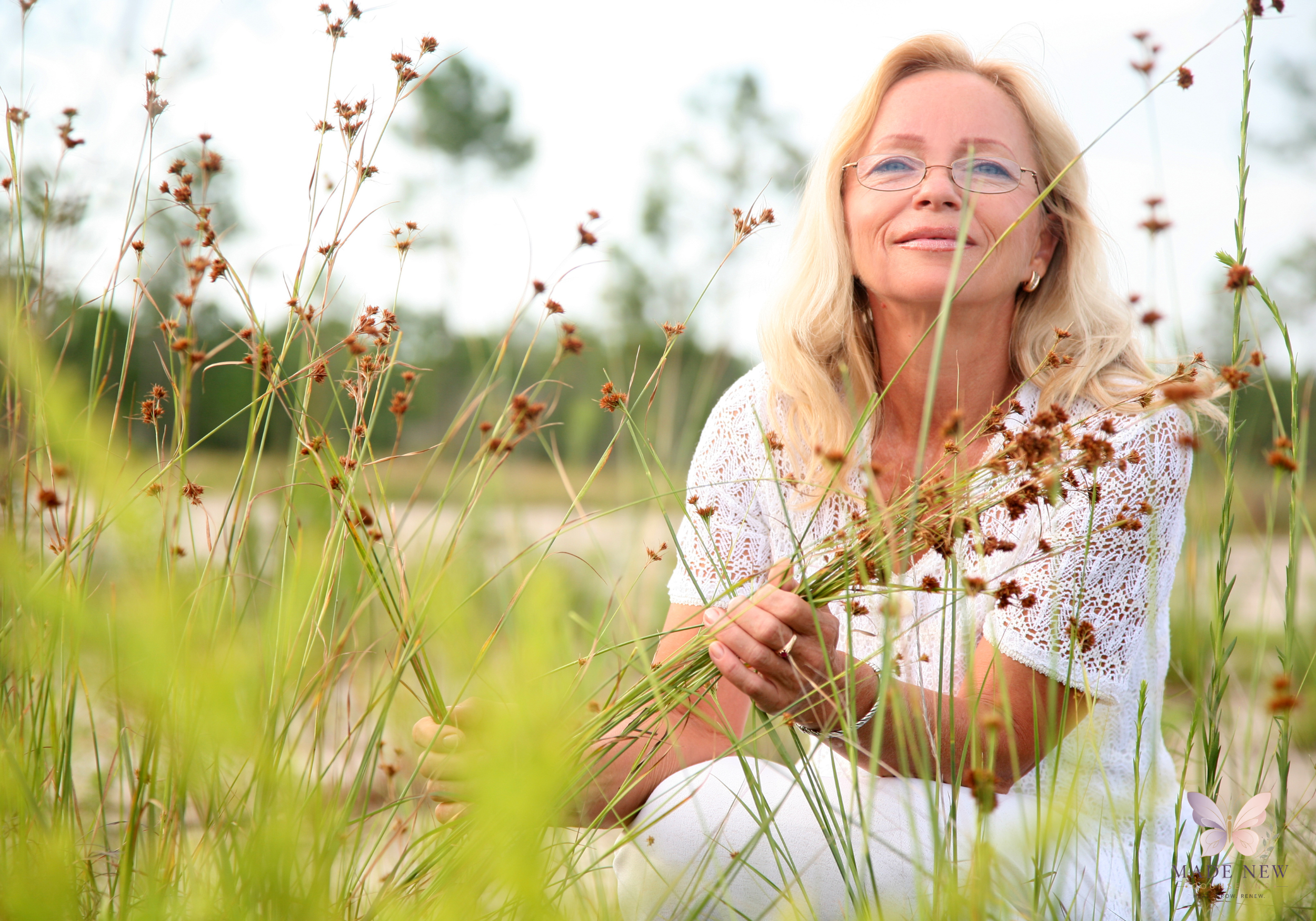 Older blonde woman with glasses sits in tall grass holding wild plants, smiling gently toward the sky.