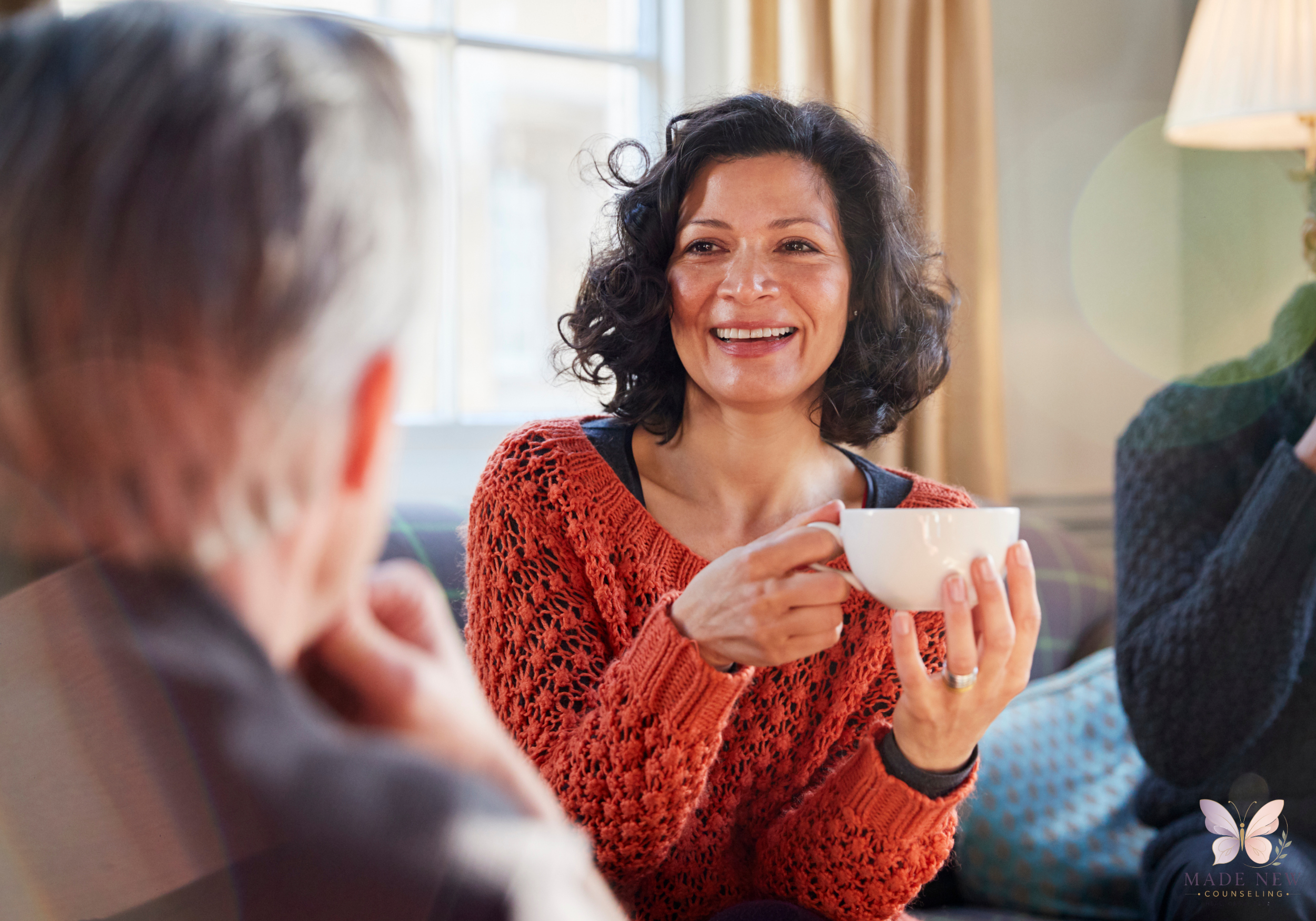 Woman in an orange sweater smiles while holding a teacup during a warm conversation indoor