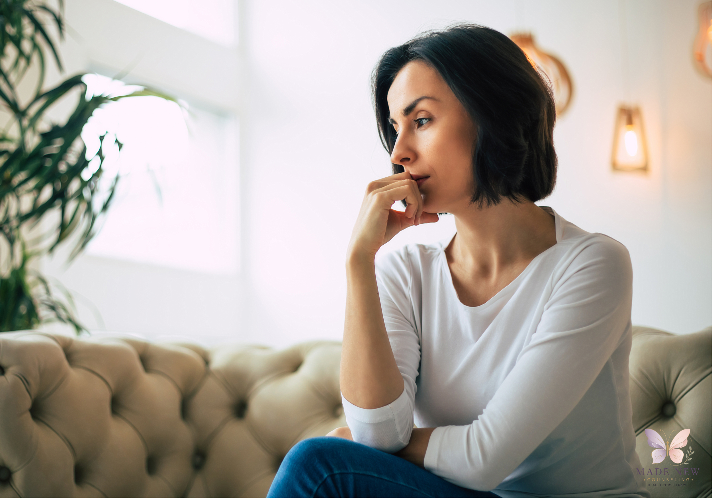 Thoughtful woman with short dark hair sits on a beige couch, hand near her mouth, gazing into the distance.