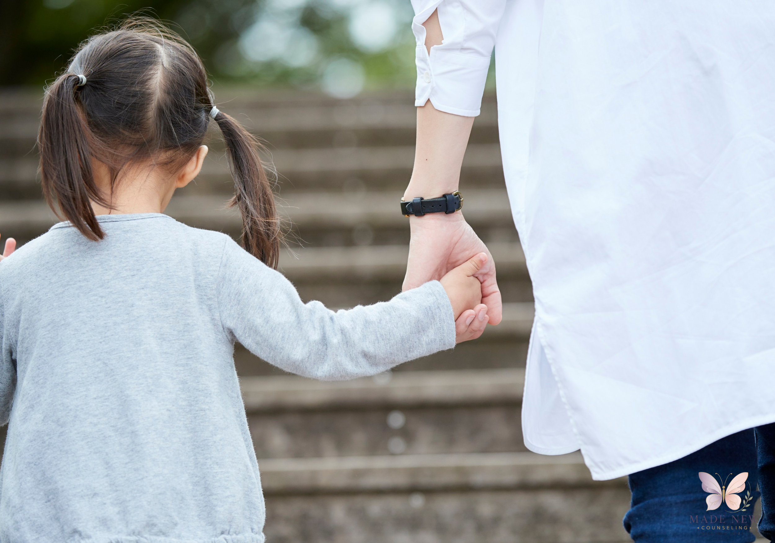Adult in a white shirt holds a young child’s hand as they walk up outdoor concrete steps together.