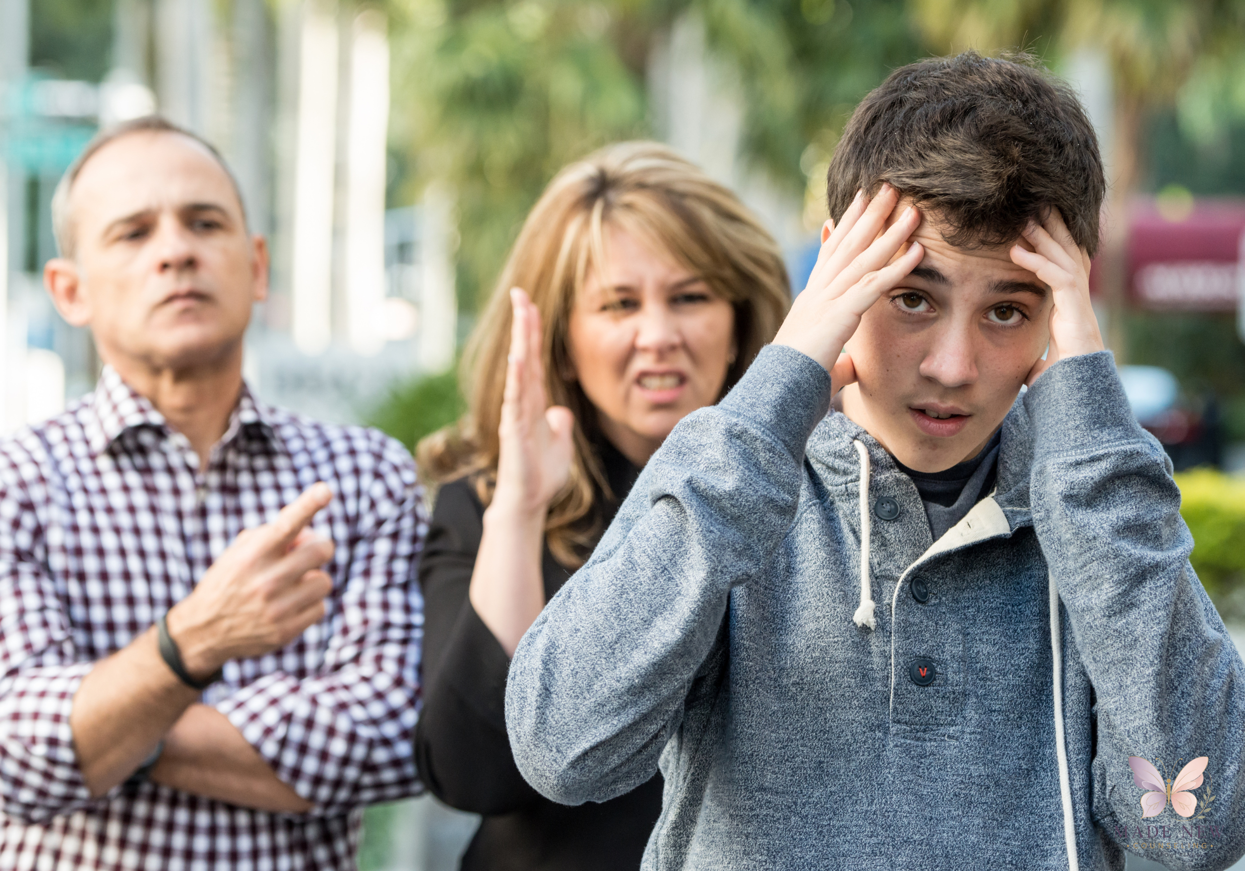 Teen covers his ears in distress while parents argue behind him outdoors.