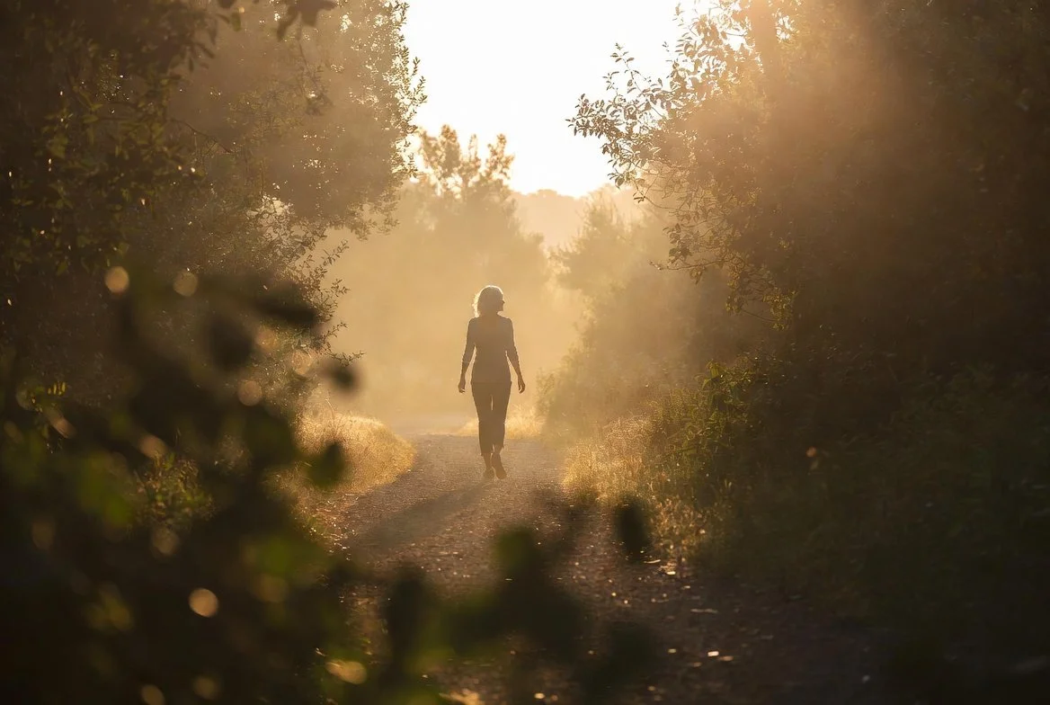 A person walking alone on a dirt path through a wooded area during sunset or sunrise, with warm golden light filtering through the trees.
