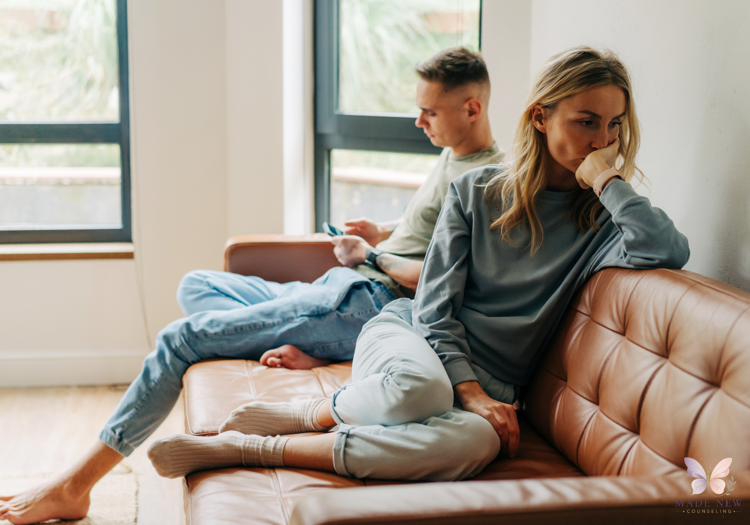Couple sits on a leather couch; the woman looks away thoughtfully while the man focuses on his phone.
