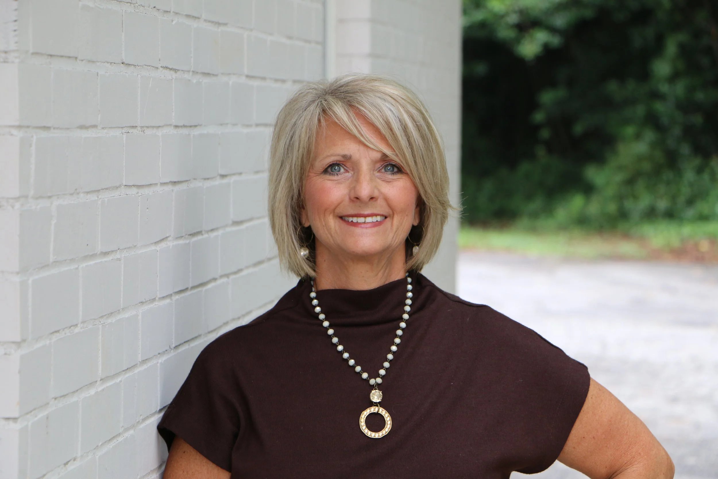 Charmion Roberts, founder of Made New Counseling, smiling outdoors in a brown turtleneck and pearl necklace against a white brick wall.