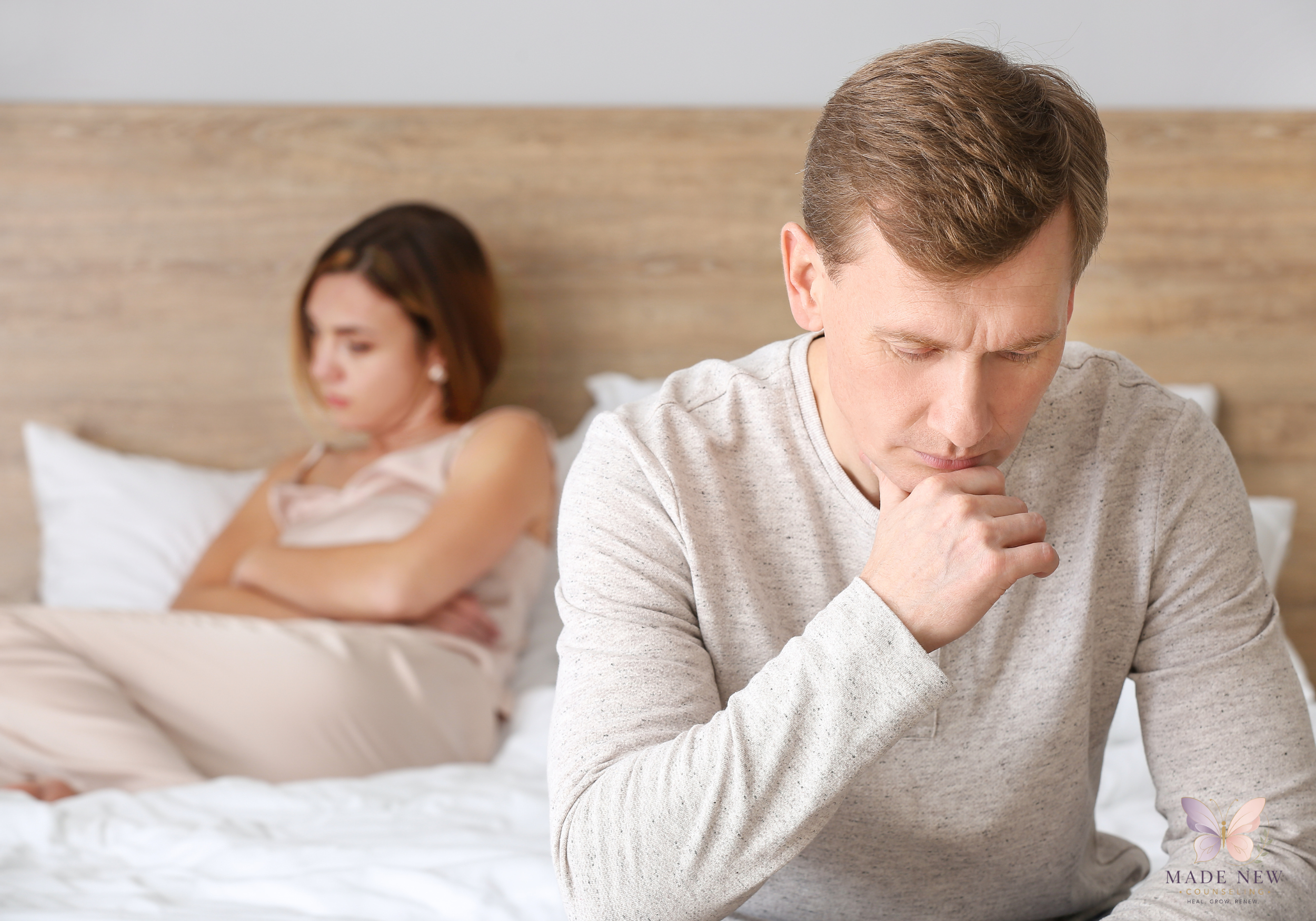 Man sits on the edge of a bed looking down thoughtfully while a woman sits behind him with arms crossed.