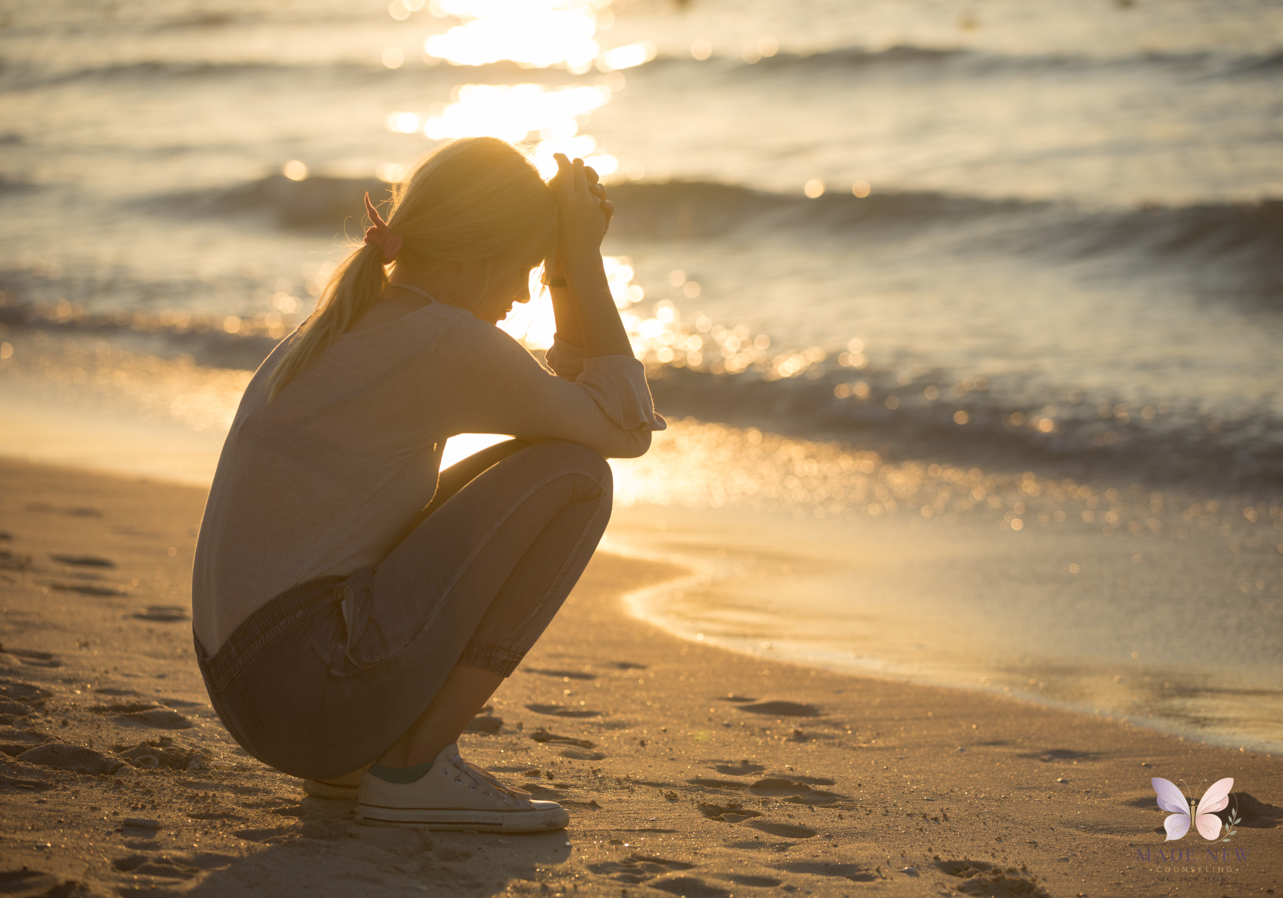 Woman crouches on a sandy beach at sunset with her head in her hands, facing the ocean waves.