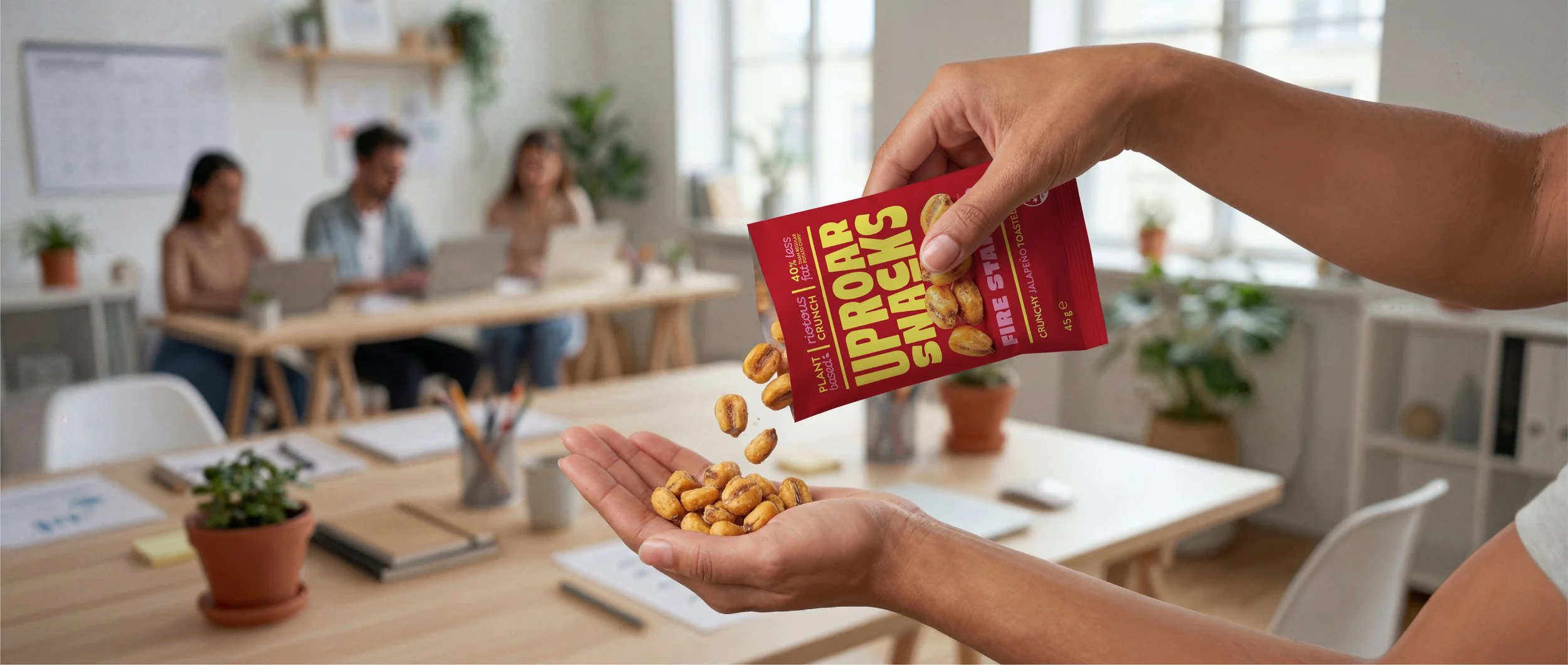 Person pouring Uppeer Snacks plant-based snack onto palm in an office meeting room with three women sitting at a table in the background.