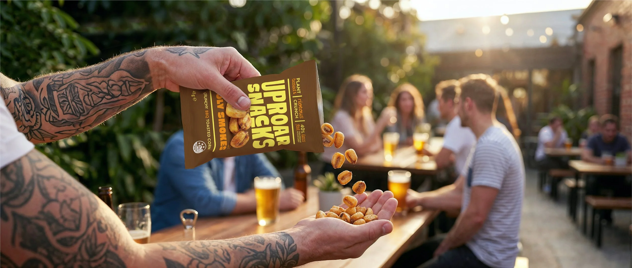 A person with tattooed arms is holding a bag of plant-based roasted snack peanuts, pouring them into their hand, while a group of people sit around outdoor tables enjoying drinks, with sunlight and greenery in the background.