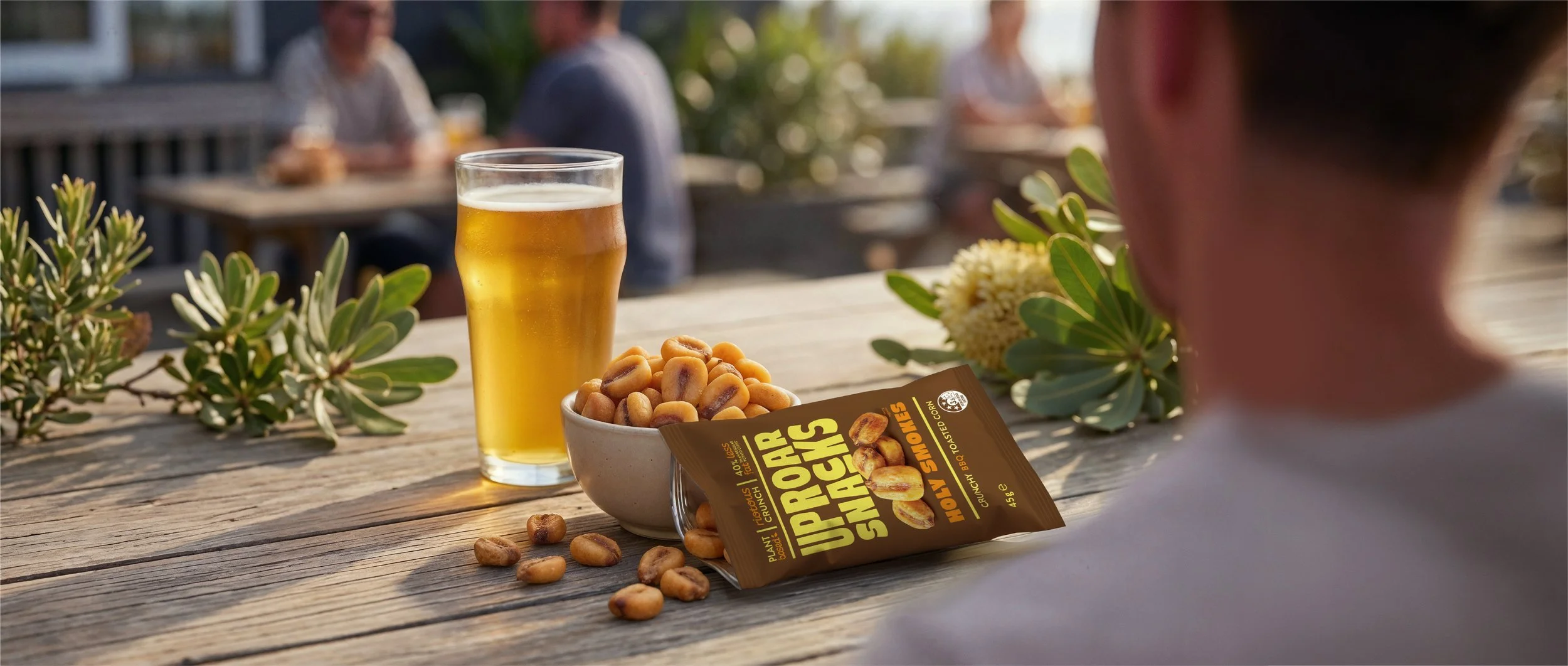 A glass of beer, a bowl of roasted peanuts, and a packet of Up For Snacks on a wooden outdoor table with people in the background.