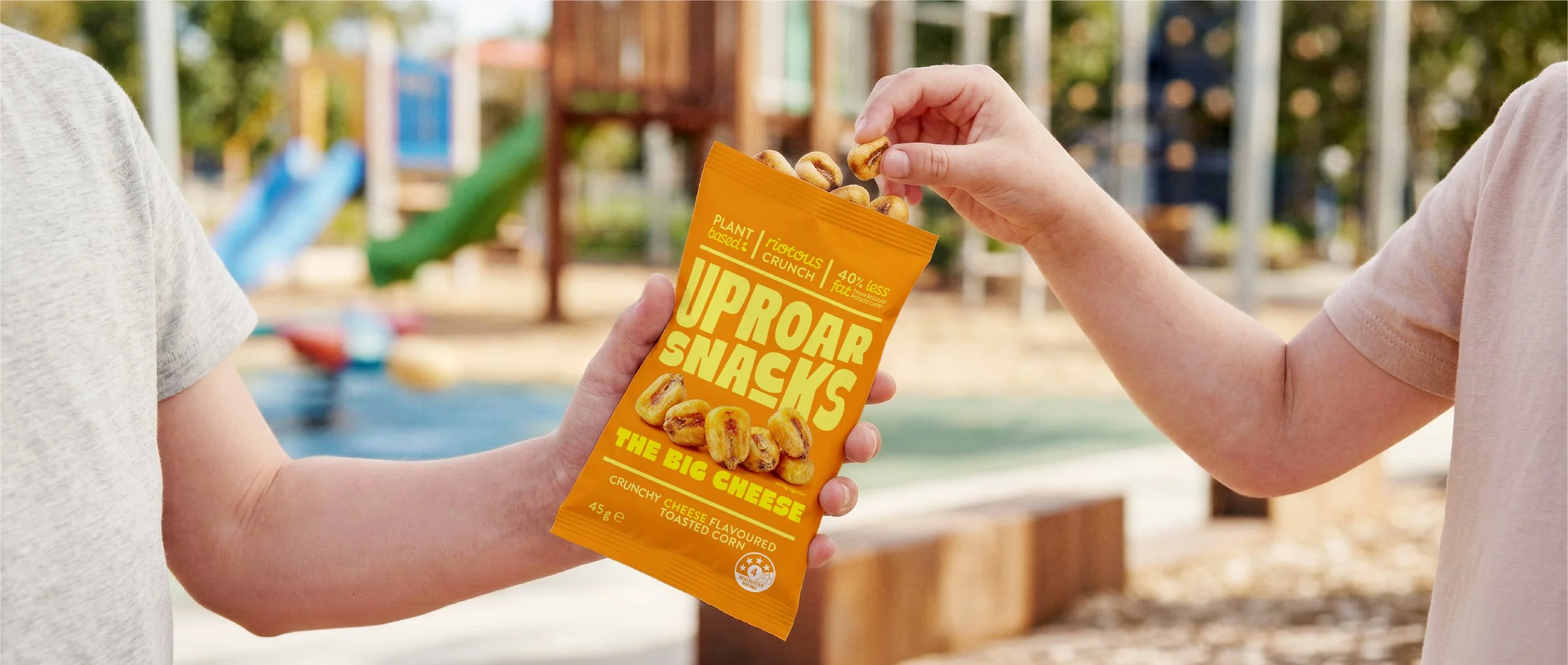 Two children exchanging a bag of Upfront Snacks, a cheese-flavored toasted corn snack, at a park.