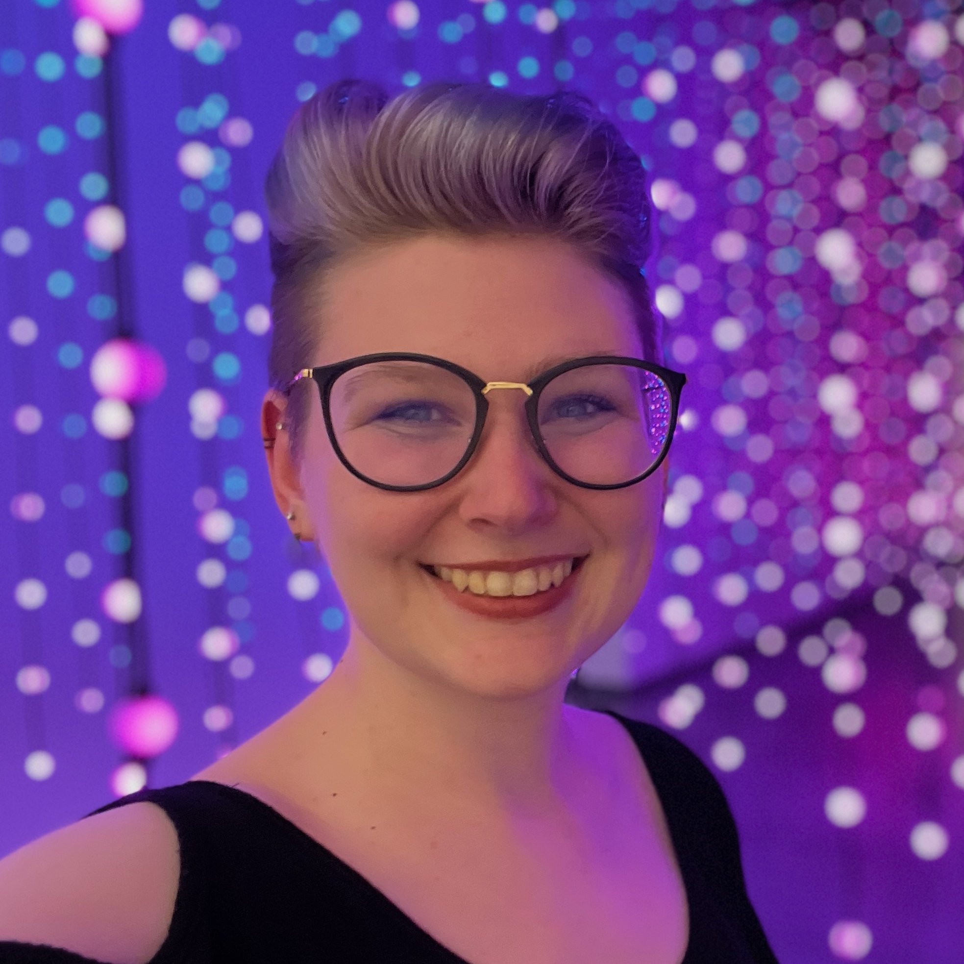 The artist Colleen smiling while wearing glasses and a black top, with a background of purple and blue bokeh lights.