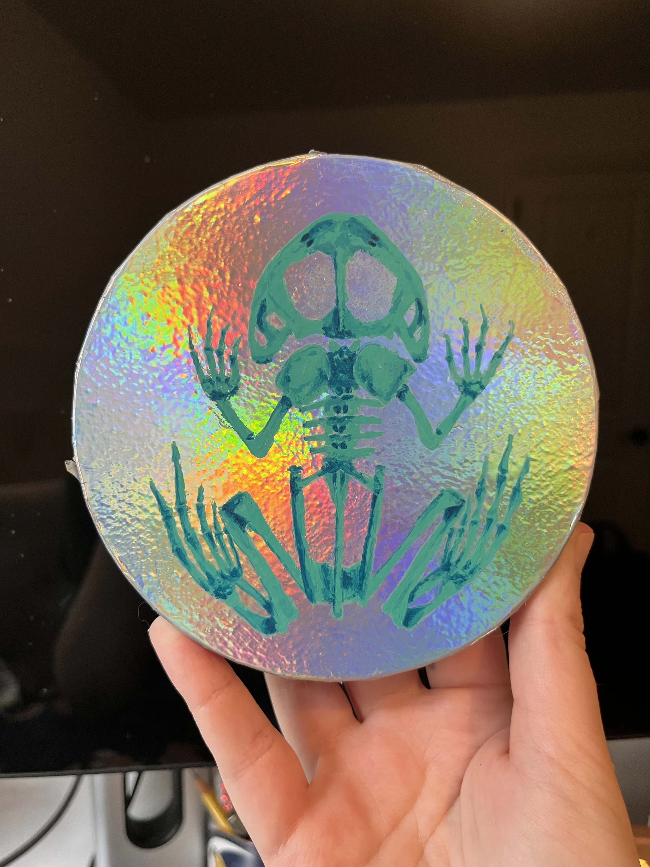 A holographic canvas features a blue frog skeleton design, with a hand holding it against a dark background.