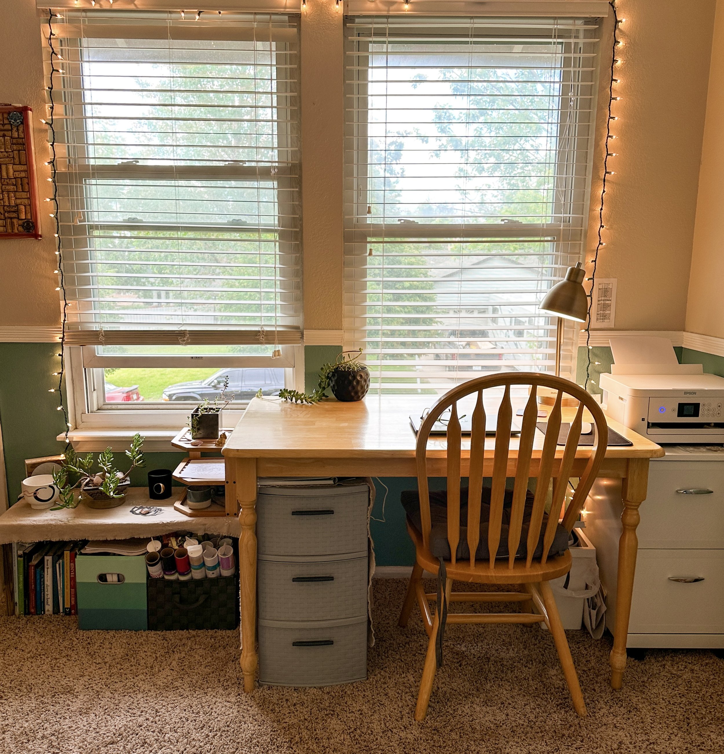 A window with blinds and string lights surrounding it. The window has a view of trees and the outside yard. There are plants, books, and craft supplies on the counter below the window, with a computer monitor, printer, and a wooden chair at the table. The room has green and beige walls, and the floor is carpeted.