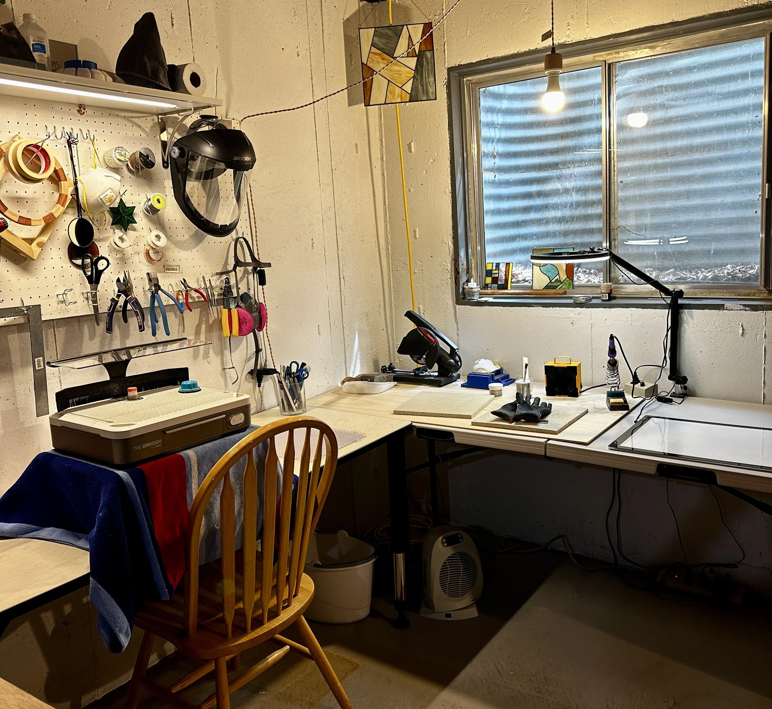 A cluttered workshop or garage with tools hanging on a pegboard, a motorcycle helmet, and a window with a corrugated metal exterior outside.