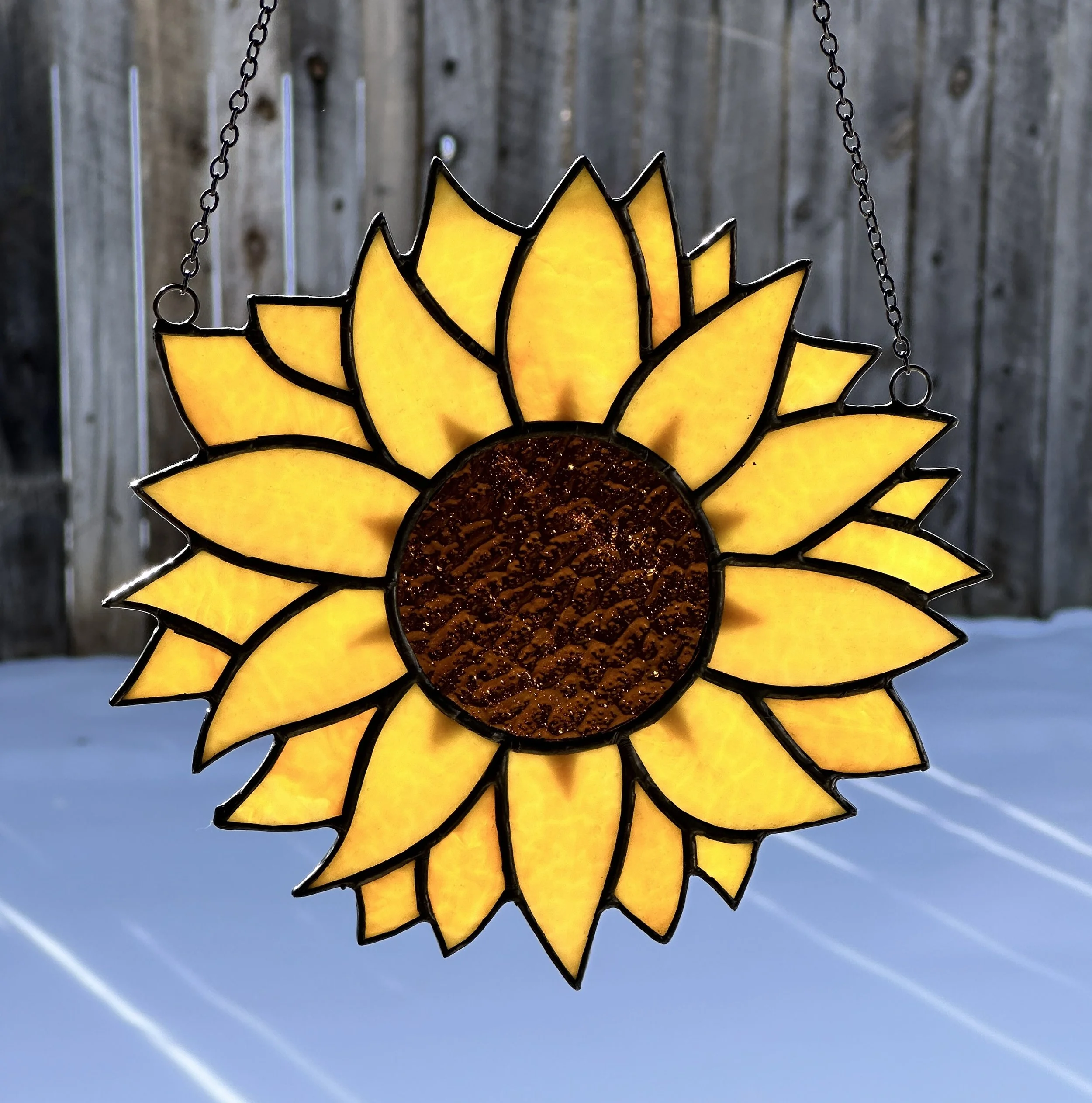 A stained glass sun-shaped decoration with yellow petals, hanging from a chain against a wooden fence and a clear blue sky with blue streaks.