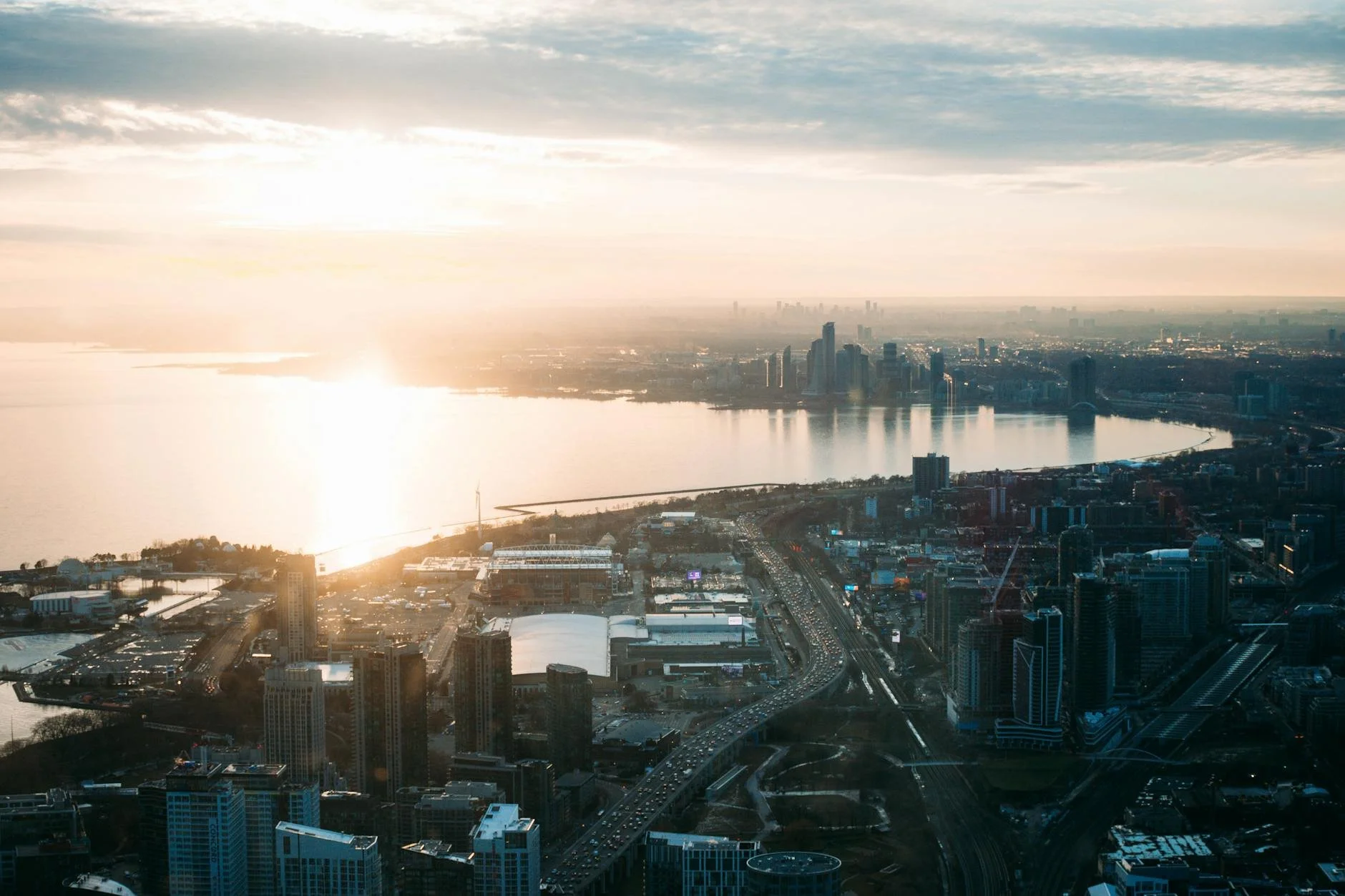 A city skyline at sunset showing tall buildings, a river, and a highway with cars.