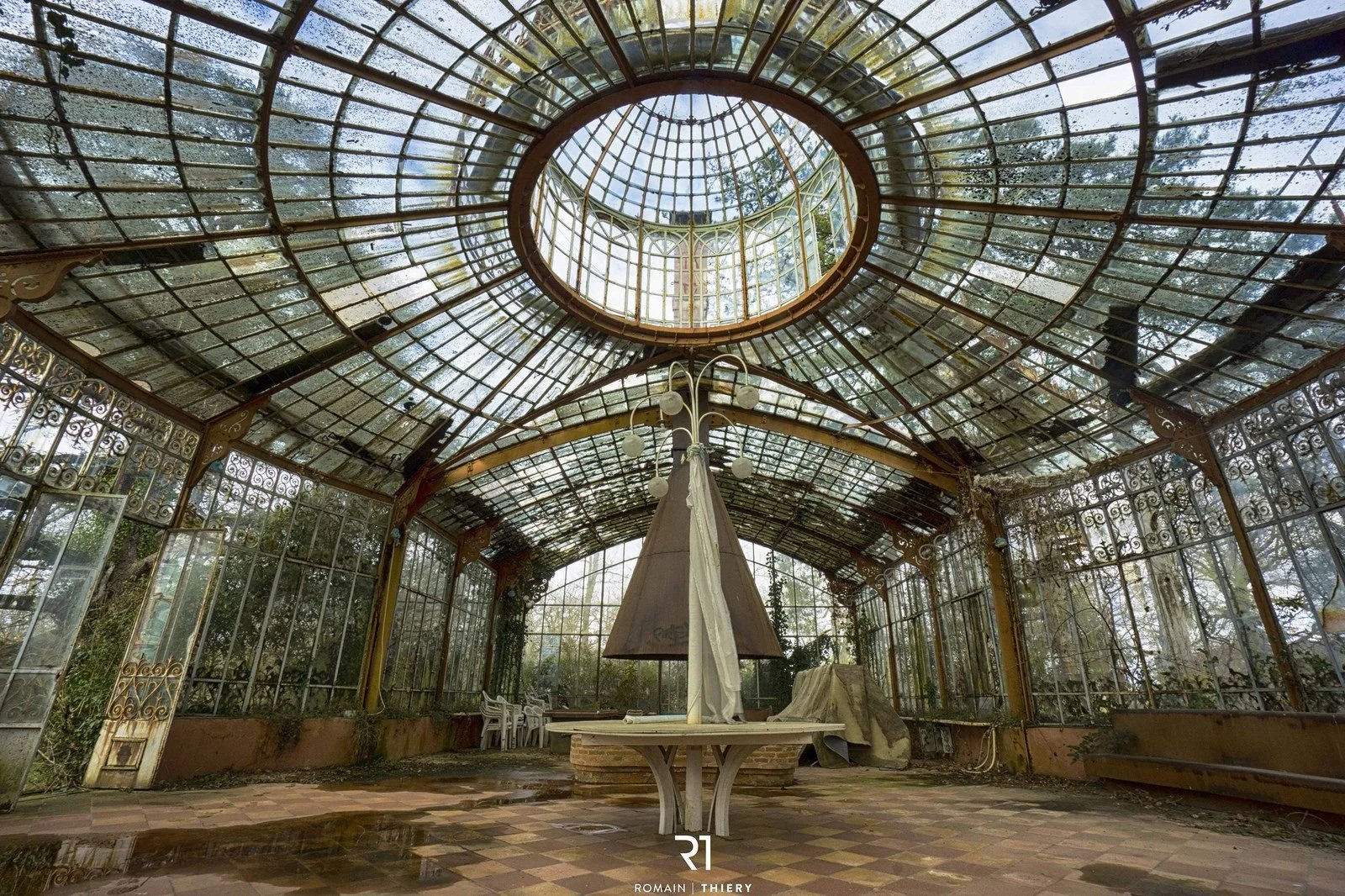 An abandoned glass greenhouse with broken windows and overgrown plants, featuring a central platform with a tent-like structure and a chandelier hanging above.