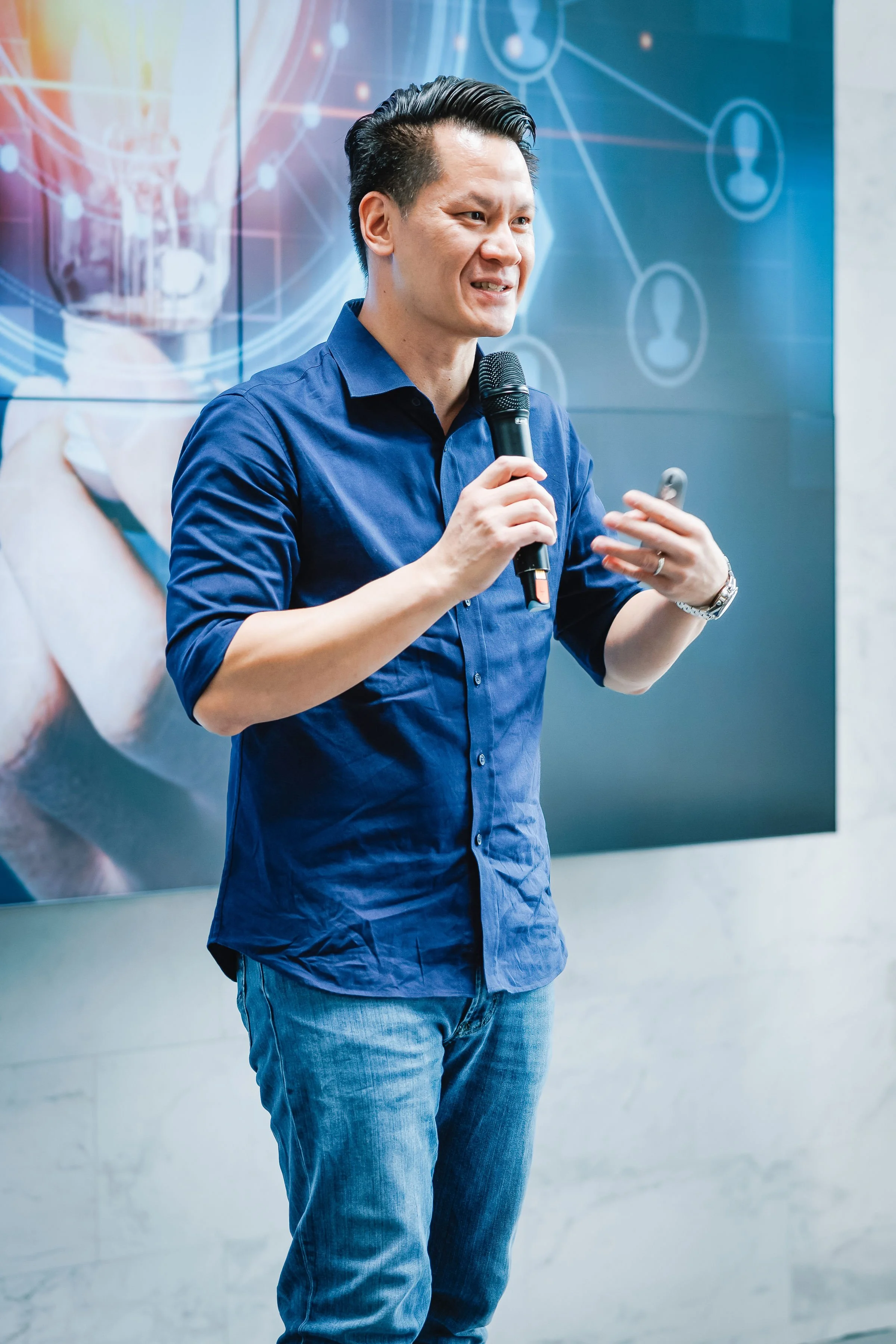 A man in a blue shirt holding a microphone and speaking, standing in front of a digital background with interconnected circles and icons.
