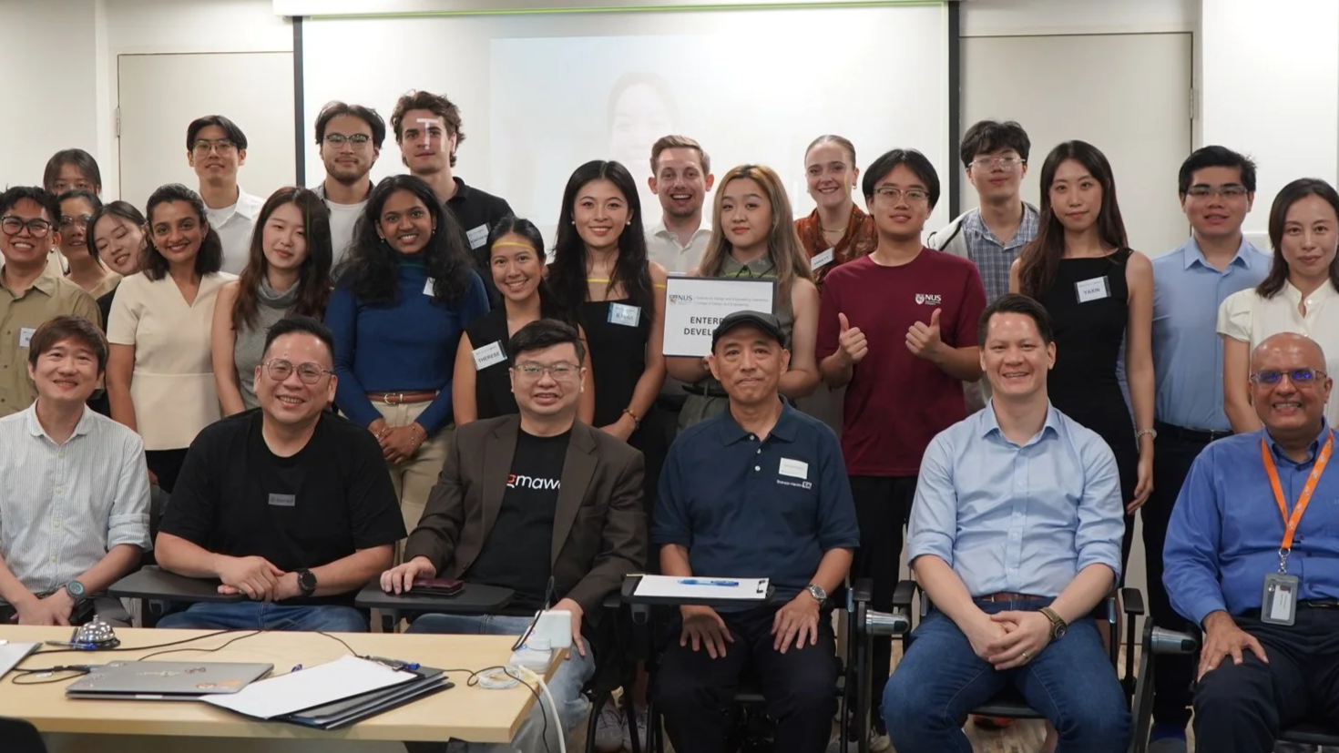 Group of young men and women, along with some older men, gathered in a classroom for a photo. Some individuals are holding a sign that reads 'ENTERPRISE DEVELOP' and wearing name tags. The group appears to be celebrating or concluding a workshop or seminar.