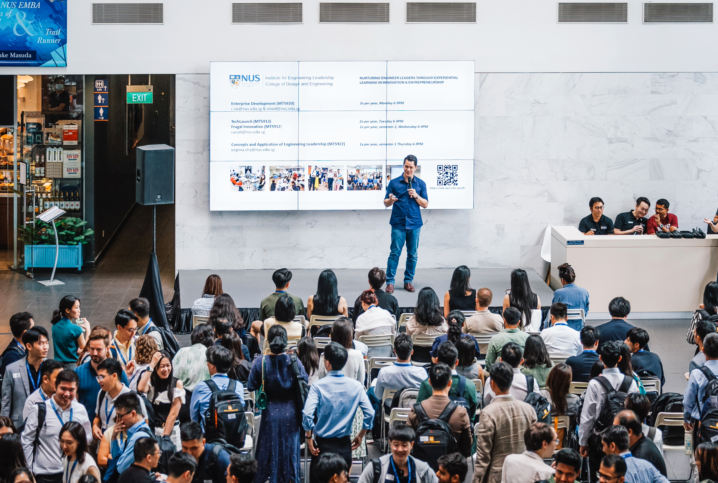 A crowded conference hall with many attendees sitting and standing, a speaker standing on stage with a microphone, large digital screens displaying presentation information about NUS EMBA and college programs, and a small group of three people seated at a table to the right of the stage.