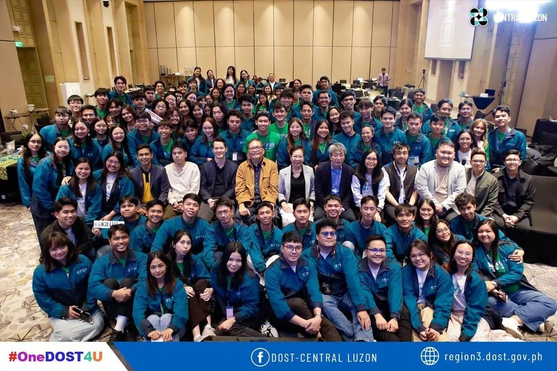 A large group of students and adults gathered in a conference room for a group photo, many wearing blue jackets with 'STTP' on them.