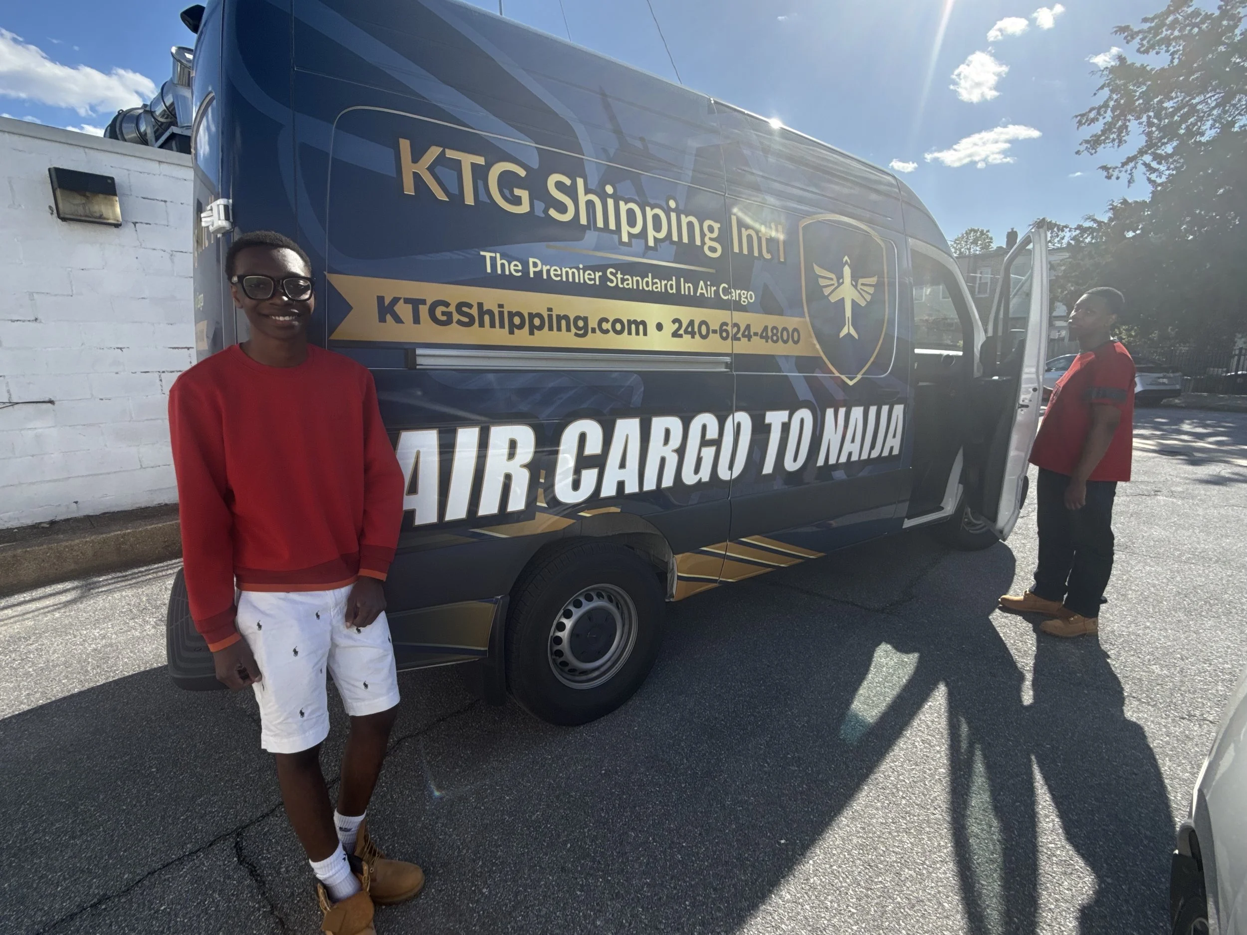 Two young men standing beside a large blue shipping van with gold and white text that reads "KTG Shipping Intl" and "AIR CARGO TO NAJJA." One is wearing a red sweater, white shorts, and glasses, and is smiling at the camera. The other is wearing a re