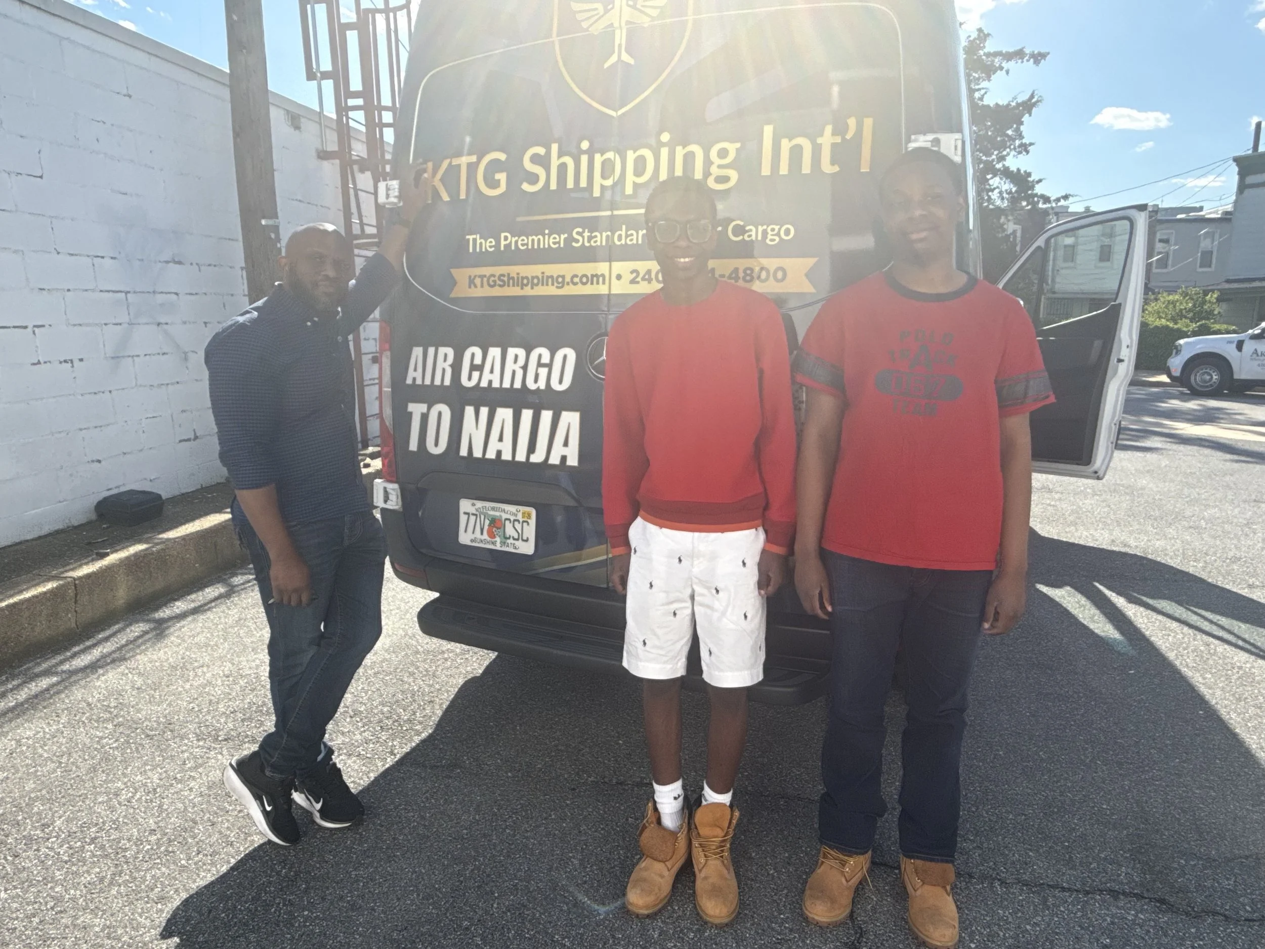 Three young men standing in front of a black KT shipping delivery vehicle, smiling. One is wearing a dark checkered shirt and jeans, another is in bright red shorts with a red shirt, and the third is in a red t-shirt and black cargo pants with Timber