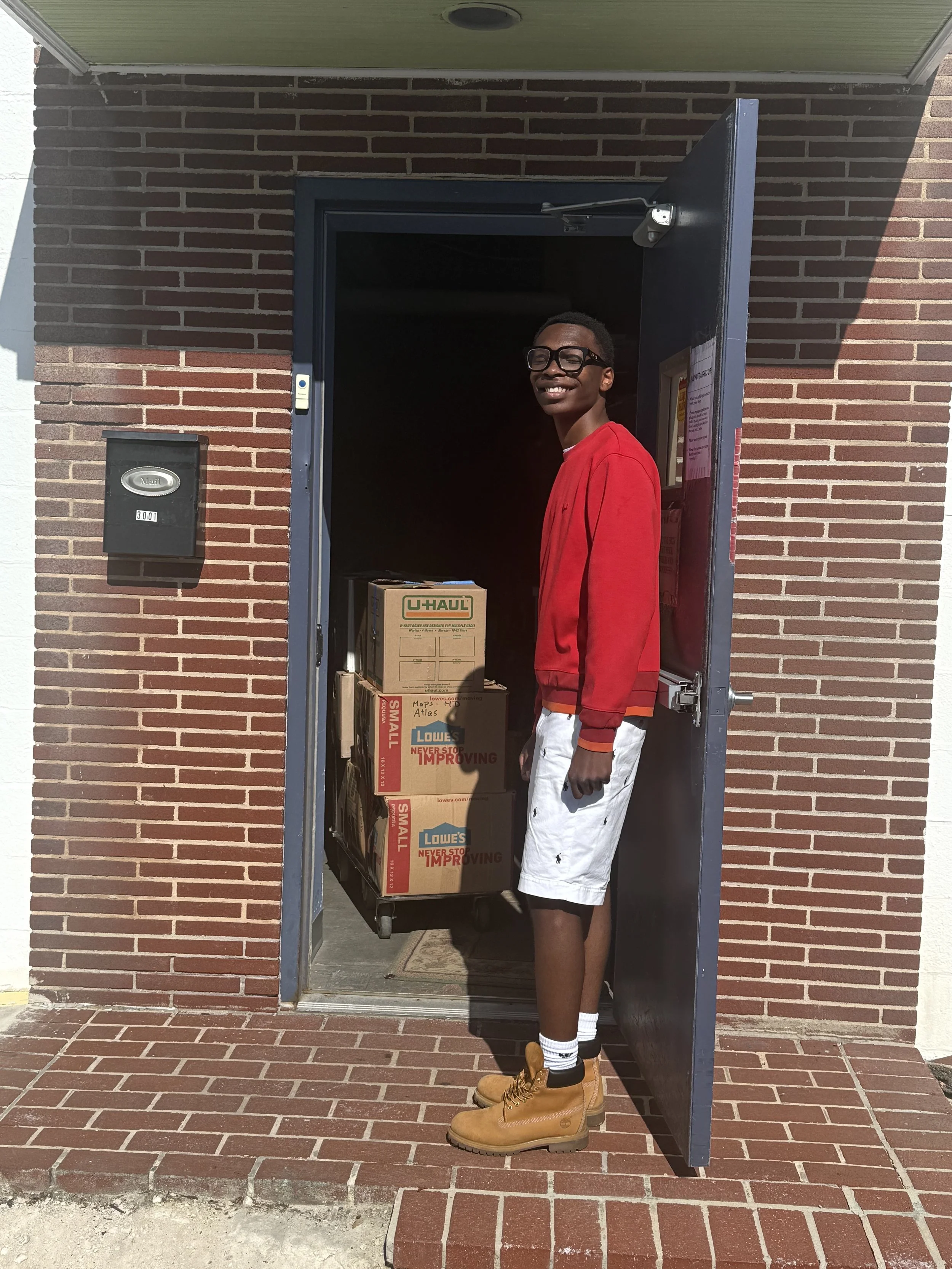A young man wearing a red jacket, white shorts, and tan work boots standing in a doorway with a partially loaded cart of boxes inside a brick building.