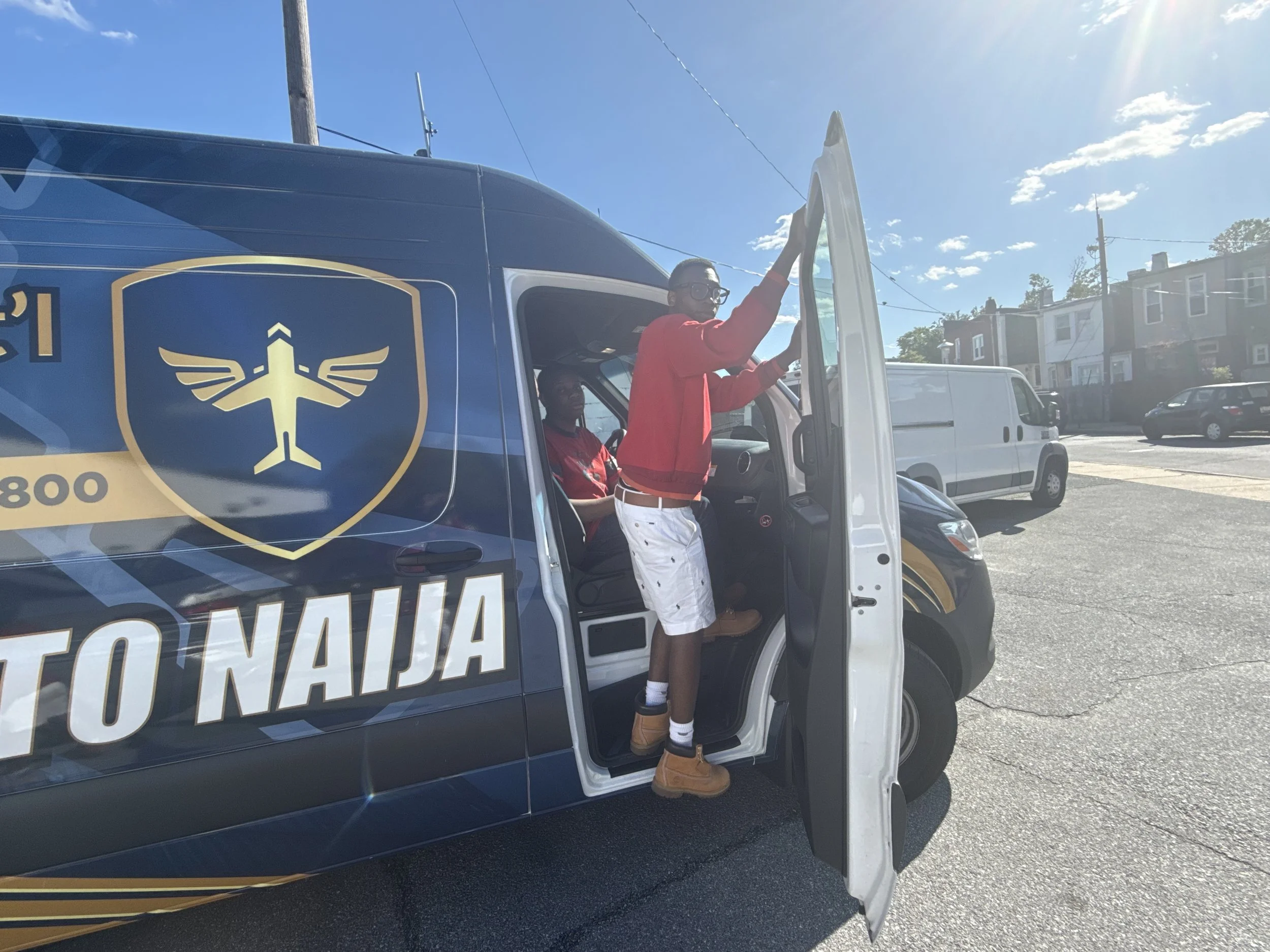 A person wearing glasses, a red jacket, white shorts with black spots, and tan boots is sitting in the driver's seat of a U.S. Postal Service mail delivery truck, while another person in a red shirt stands outside and waves.