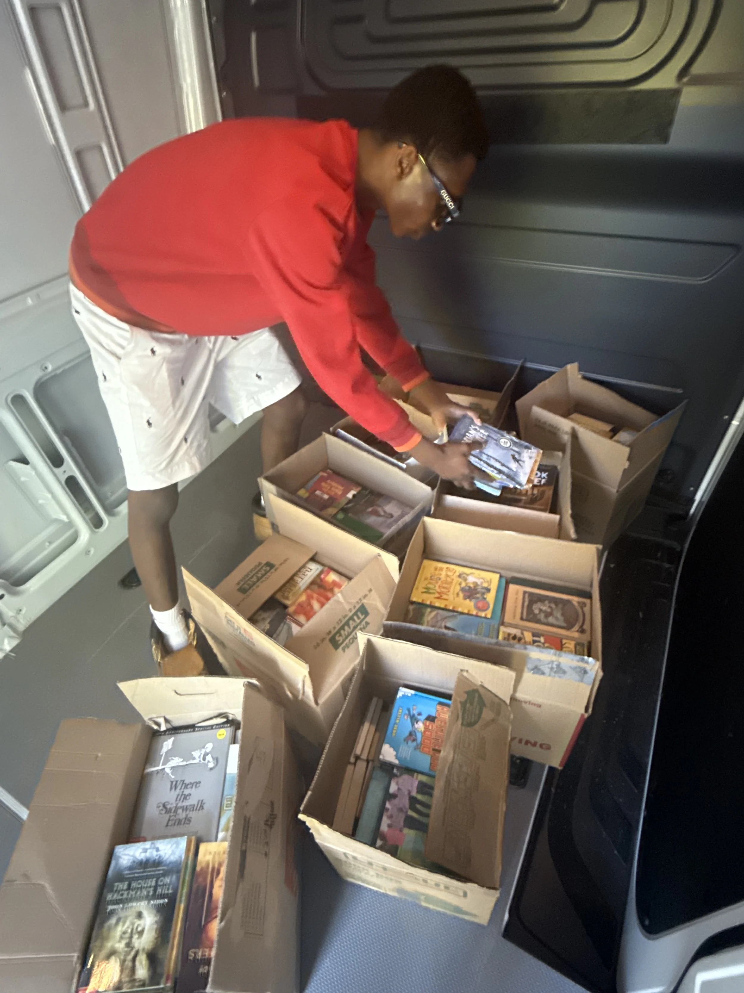 A person wearing a red jacket, white shorts, and black shoes is organizing or arranging books and magazines in cardboard boxes inside what appears to be a vehicle or storage area.