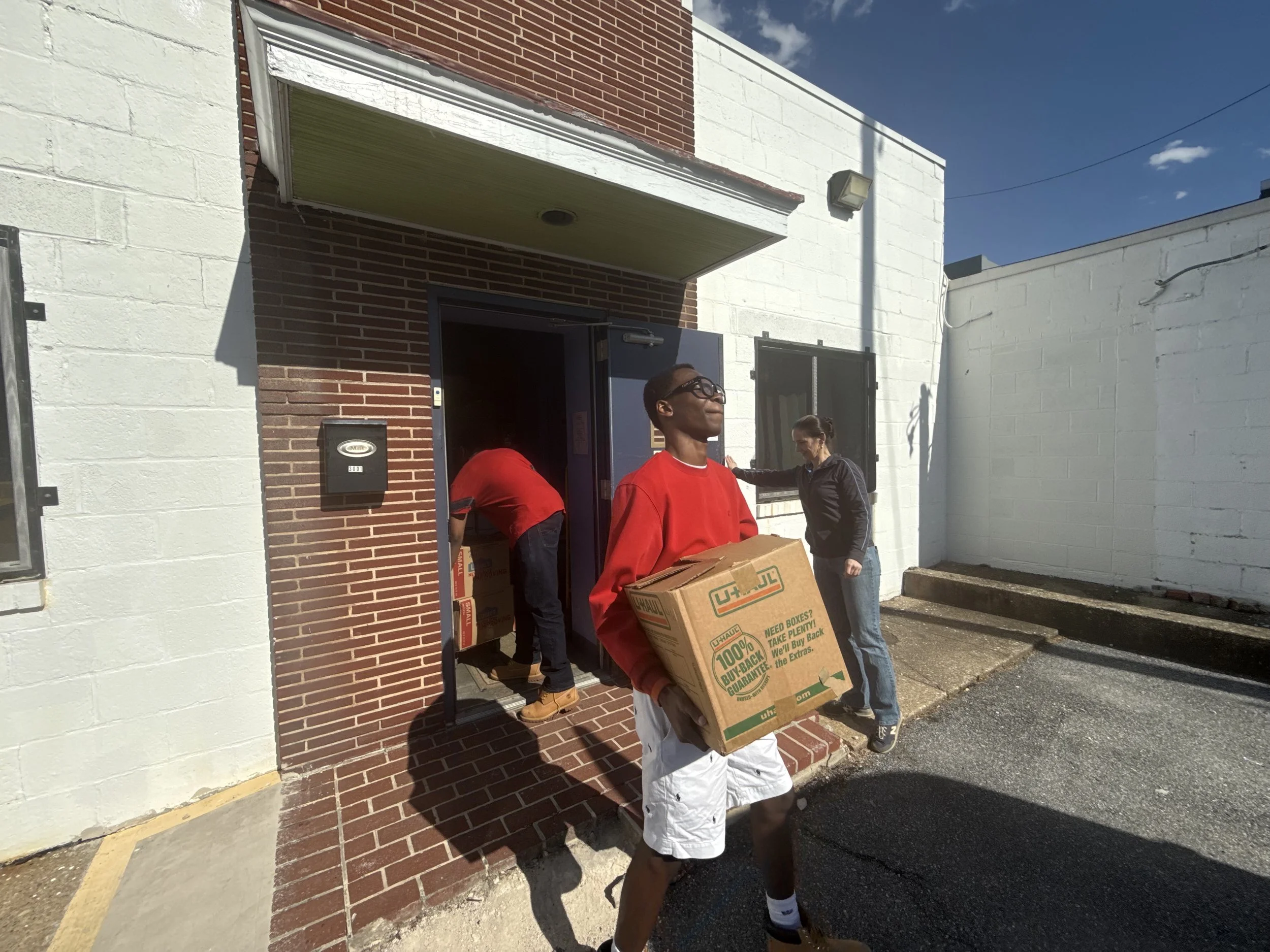 Young man carrying a cardboard box outside a building with brick and white walls, alongside a woman, on a sunny day.