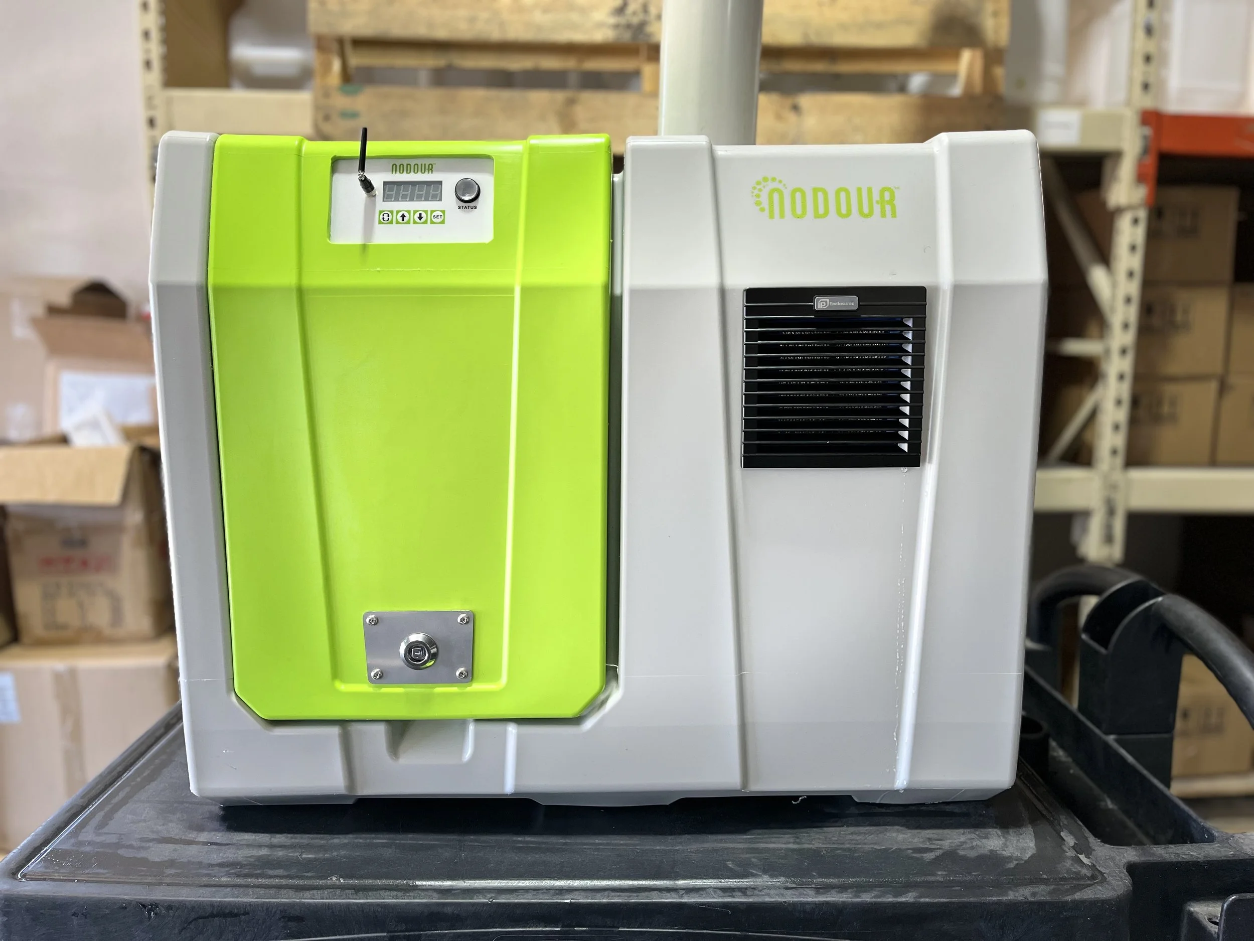 A close-up of a green and white medical device with a digital display, buttons, and an antenna, on a black surface in a warehouse setting with cardboard boxes and shelving in the background.