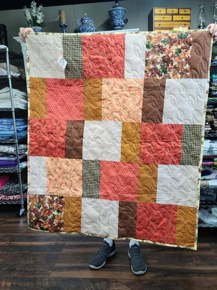 Person holding a patchwork quilt with various colored fabric squares in a store with shelves of fabric and decorative items in the background.