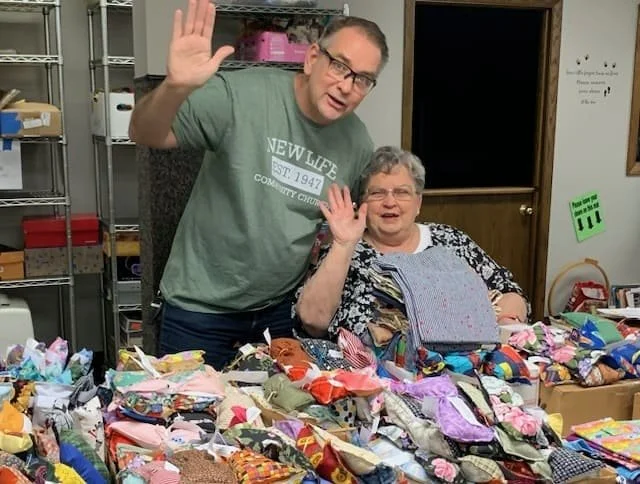 A man and woman sitting behind a table filled with handmade fabric items, both waving at the camera and smiling.