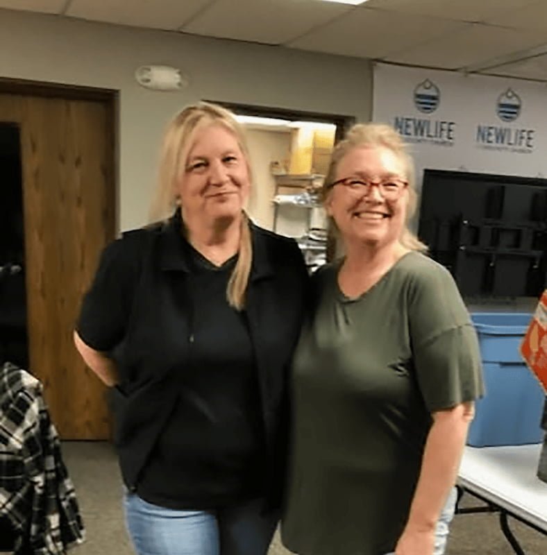Two smiling women standing indoors, one in a black shirt and the other in a green shirt, with a room that has shelves and a sign in the background.